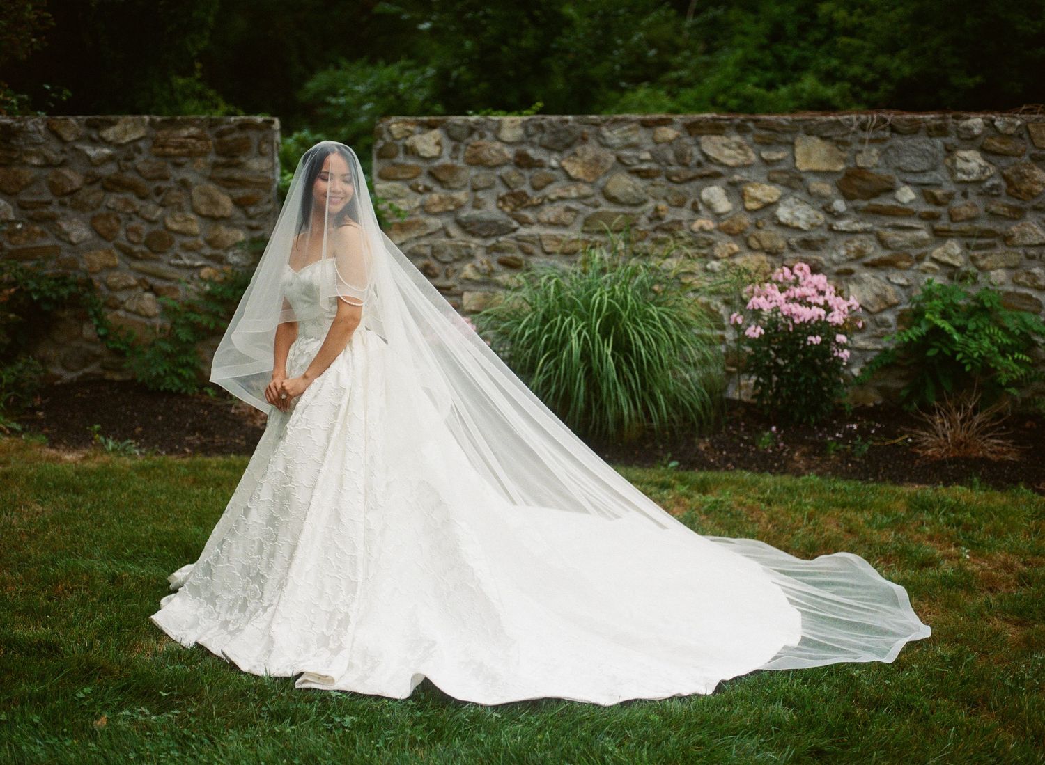 Bride in elegant lace ball gown with long cathedral veil poses on green lawn before stone wall.