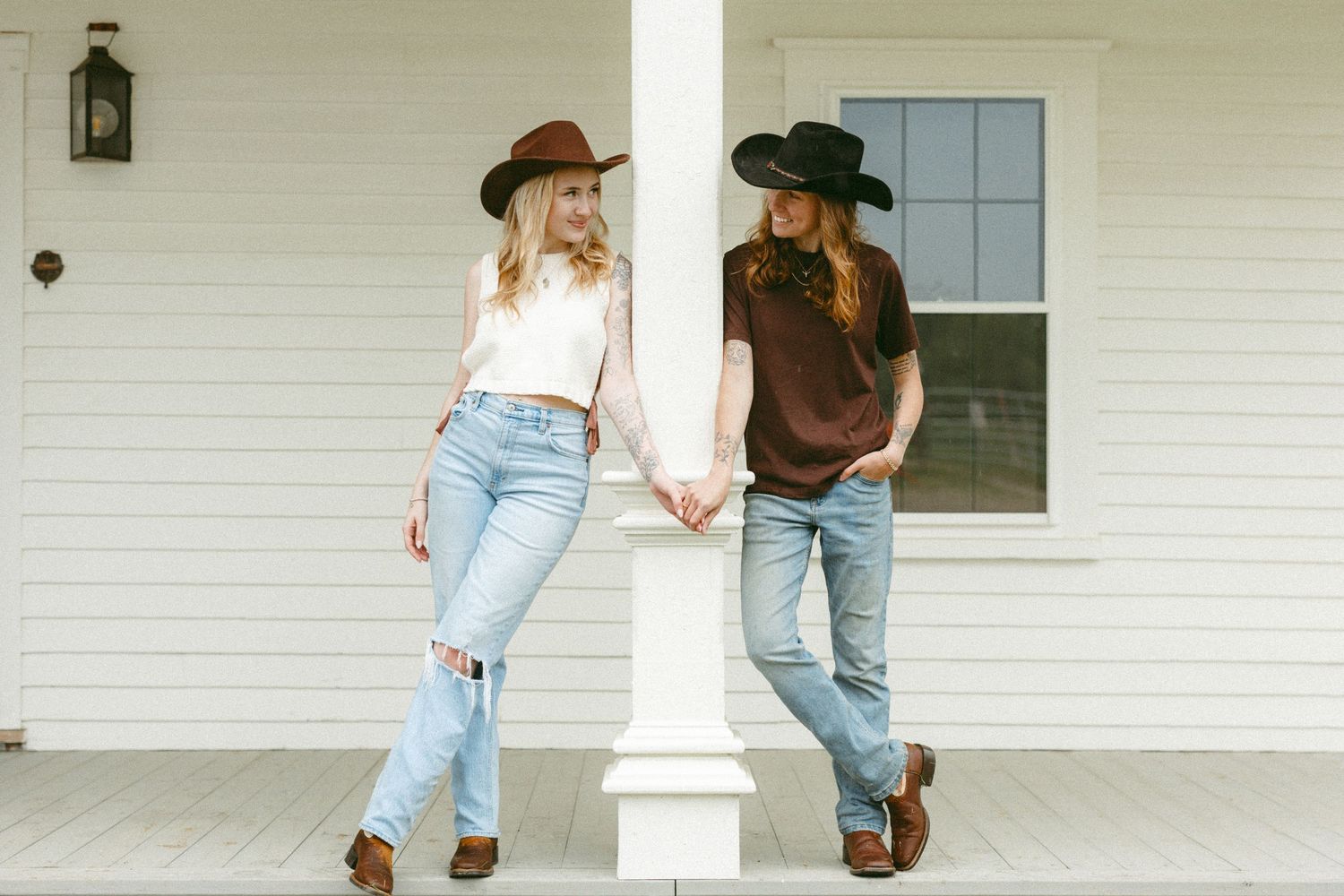 Two women in cowboy hats and jeans holding hands on a white farmhouse porch.