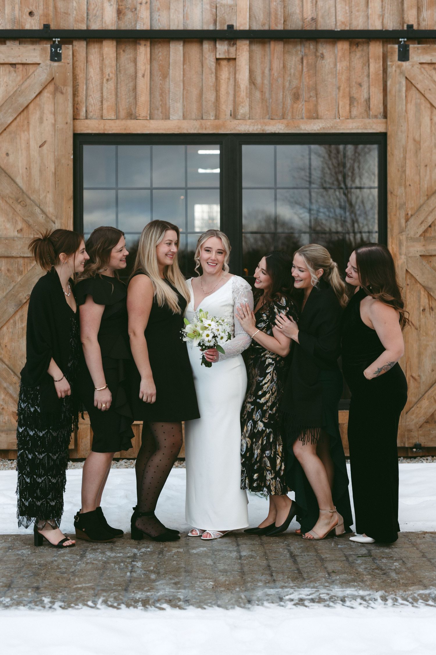Wedding party in black dresses and white gown pose creatively in front of rustic barn doors in winter setting.