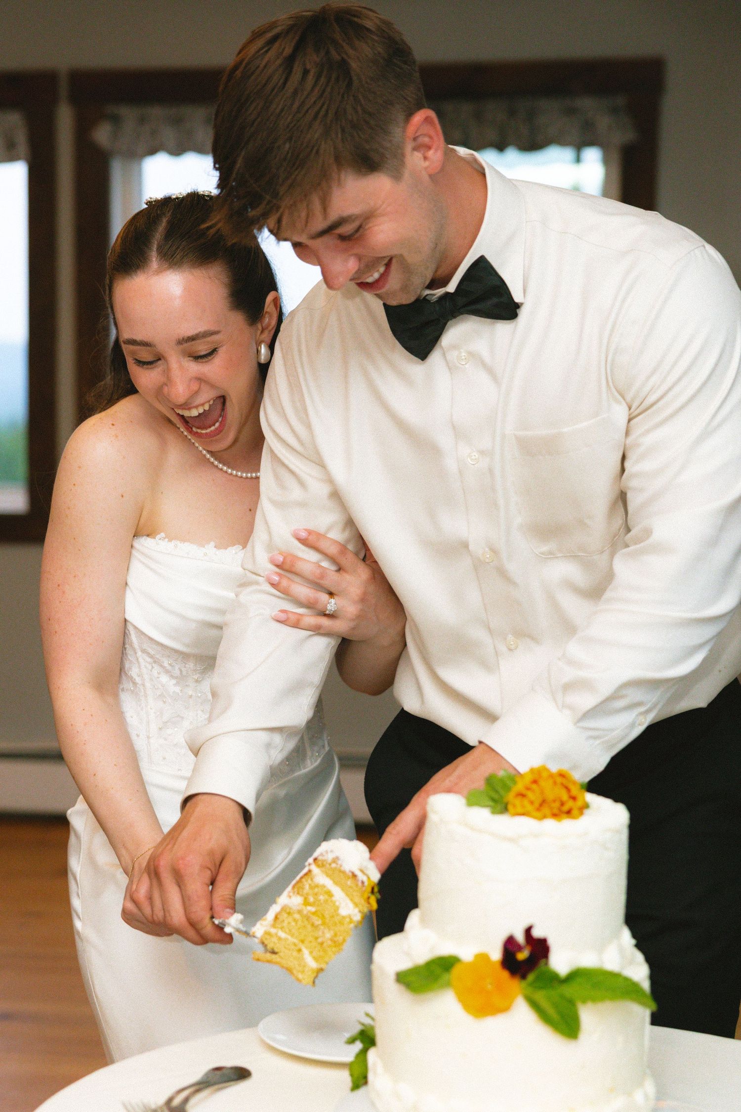 Laughing bride and groom cut their white tiered wedding cake together at their reception.