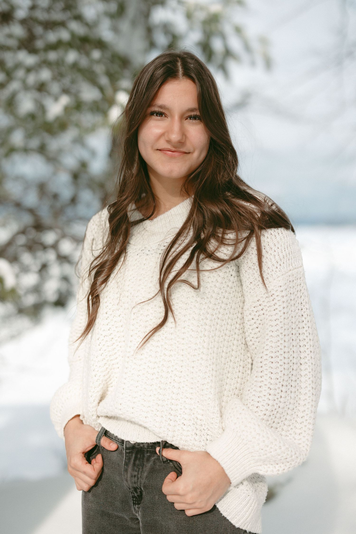 A person wearing a cozy white knit sweater and dark jeans poses outdoors against a bright winter background.