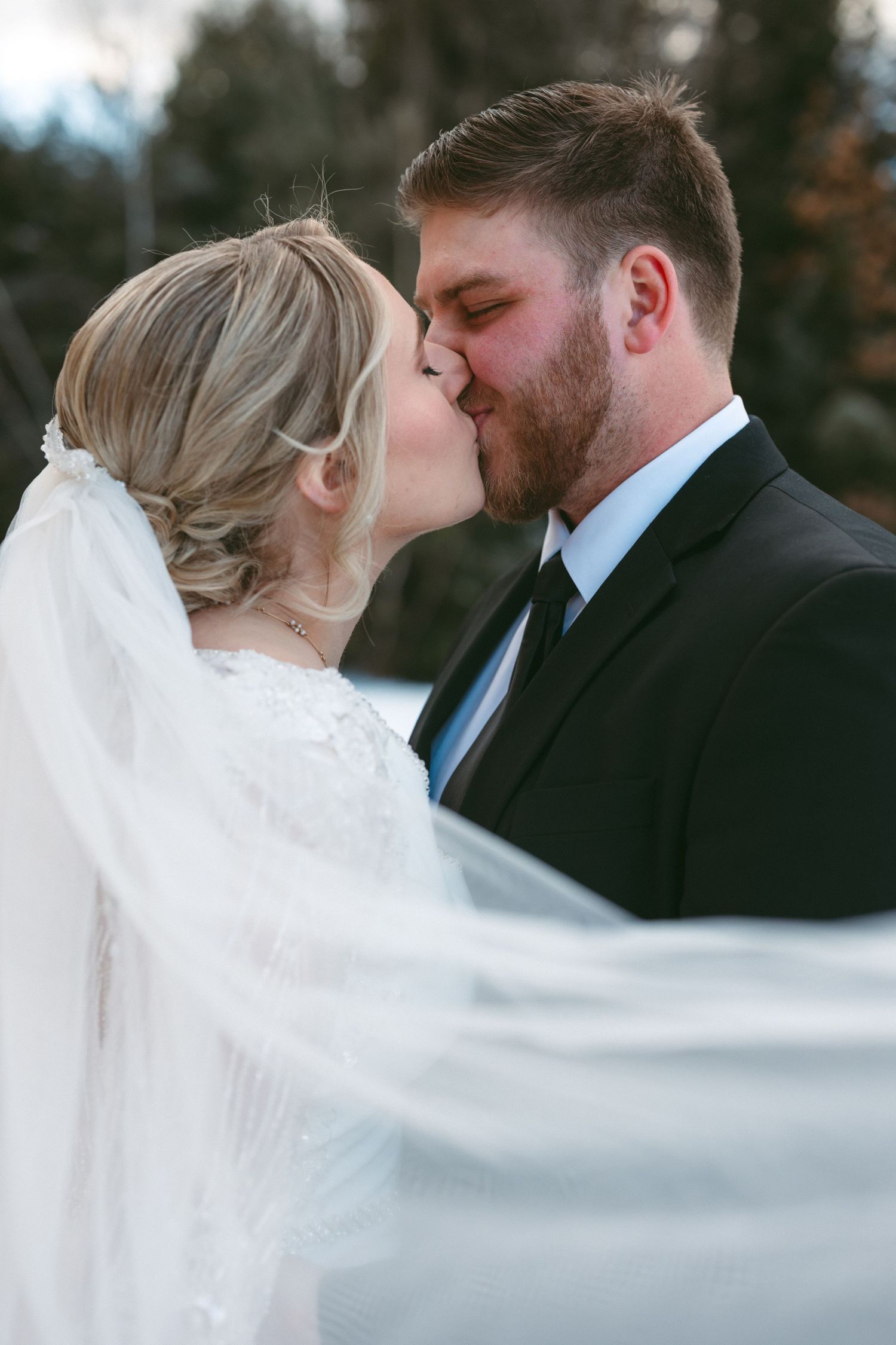 A bride and groom share an intimate kiss outdoors while her white veil flows dramatically in the wind.