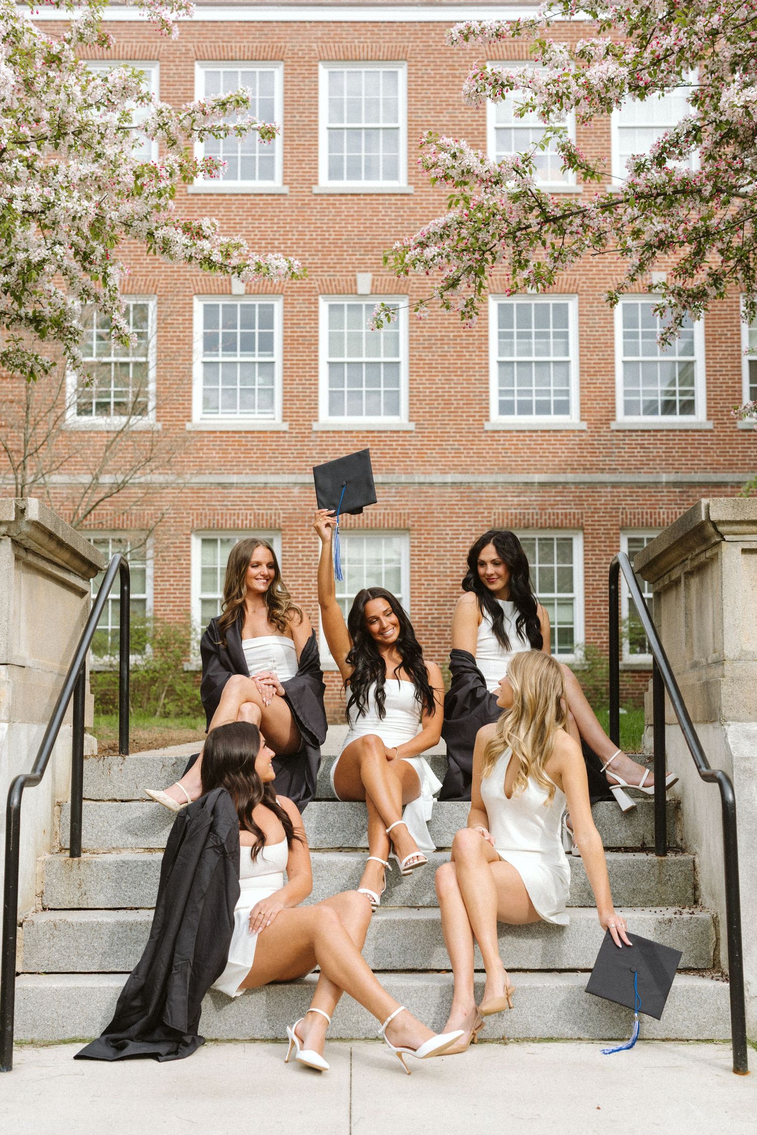 Five college graduates in white dresses and black gowns celebrate on brick building steps with graduation caps.