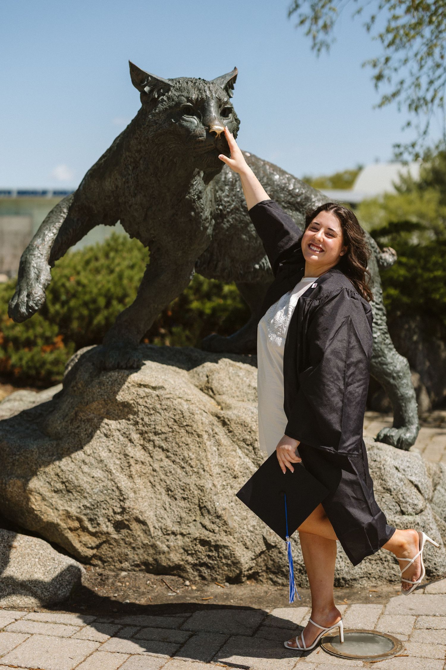 A smiling graduate in cap and gown poses beside a wildcat bronze statue outdoors on a sunny day.