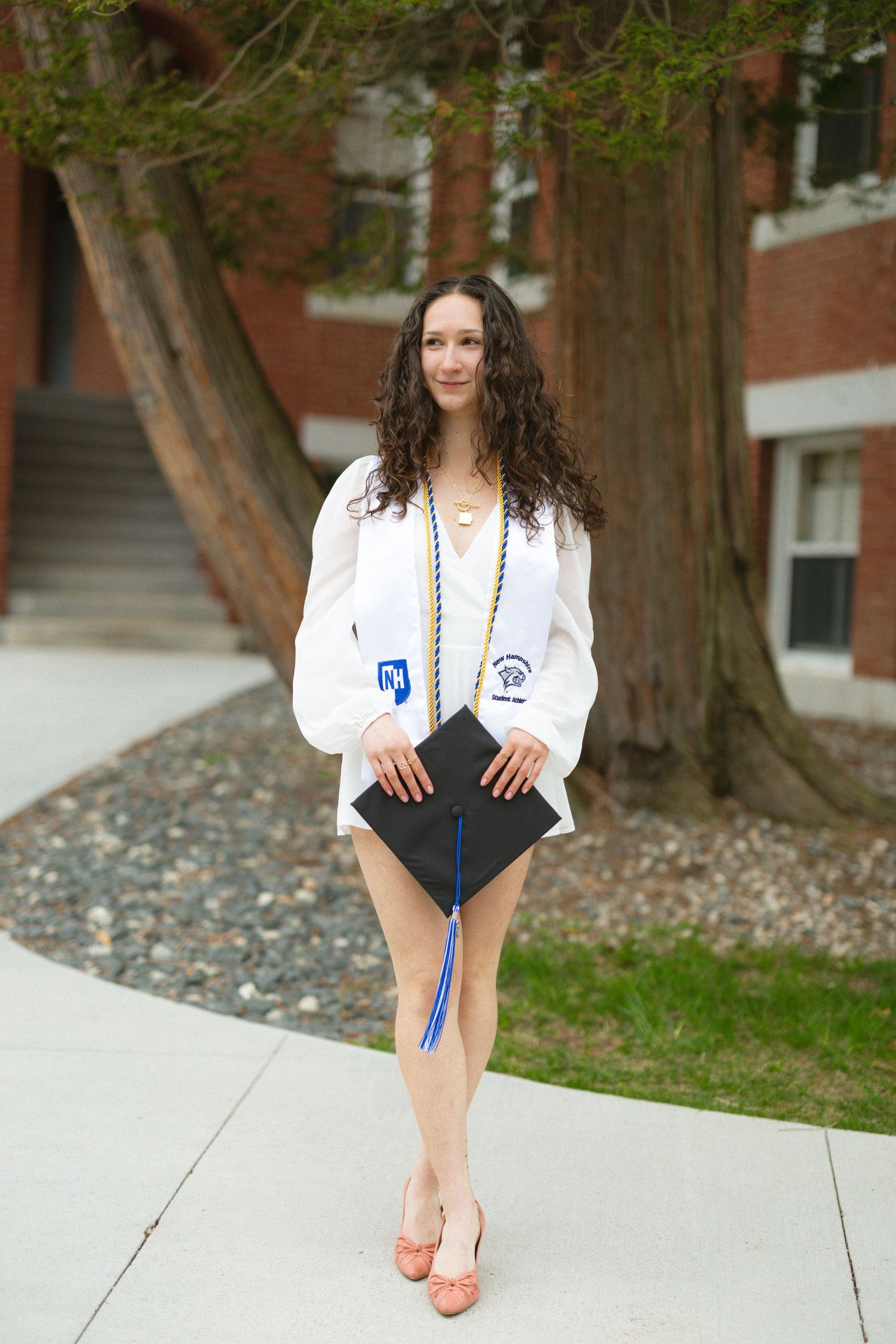 Young woman in white graduation gown holding cap, wearing coral heels, posing on campus sidewalk near brick building.