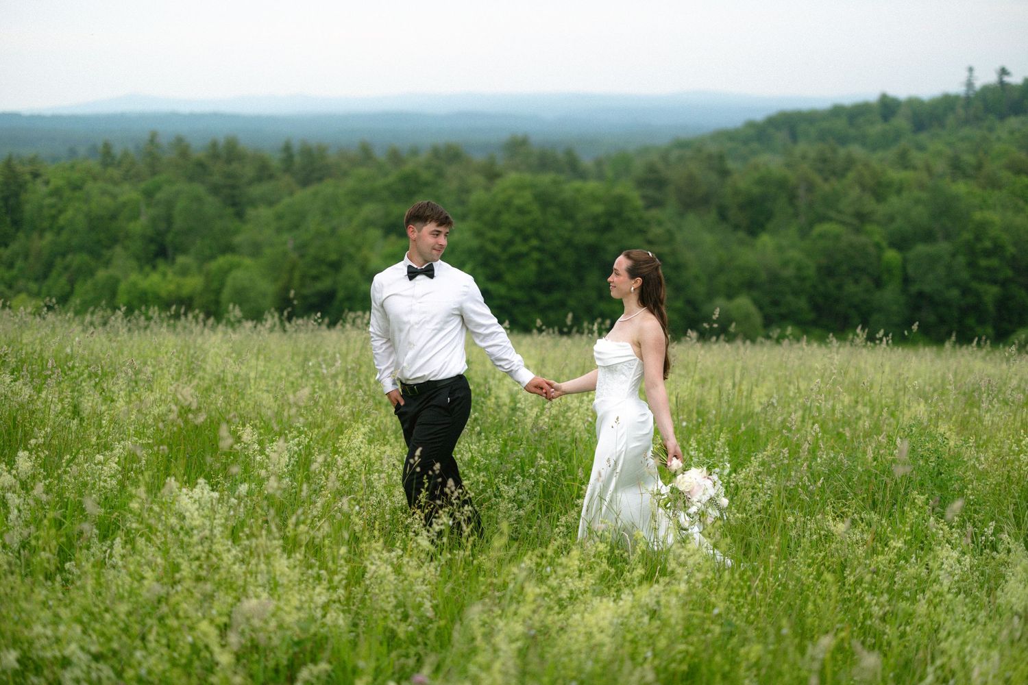Bride and groom holding hands walking through a lush green meadow with forest hills in the background.