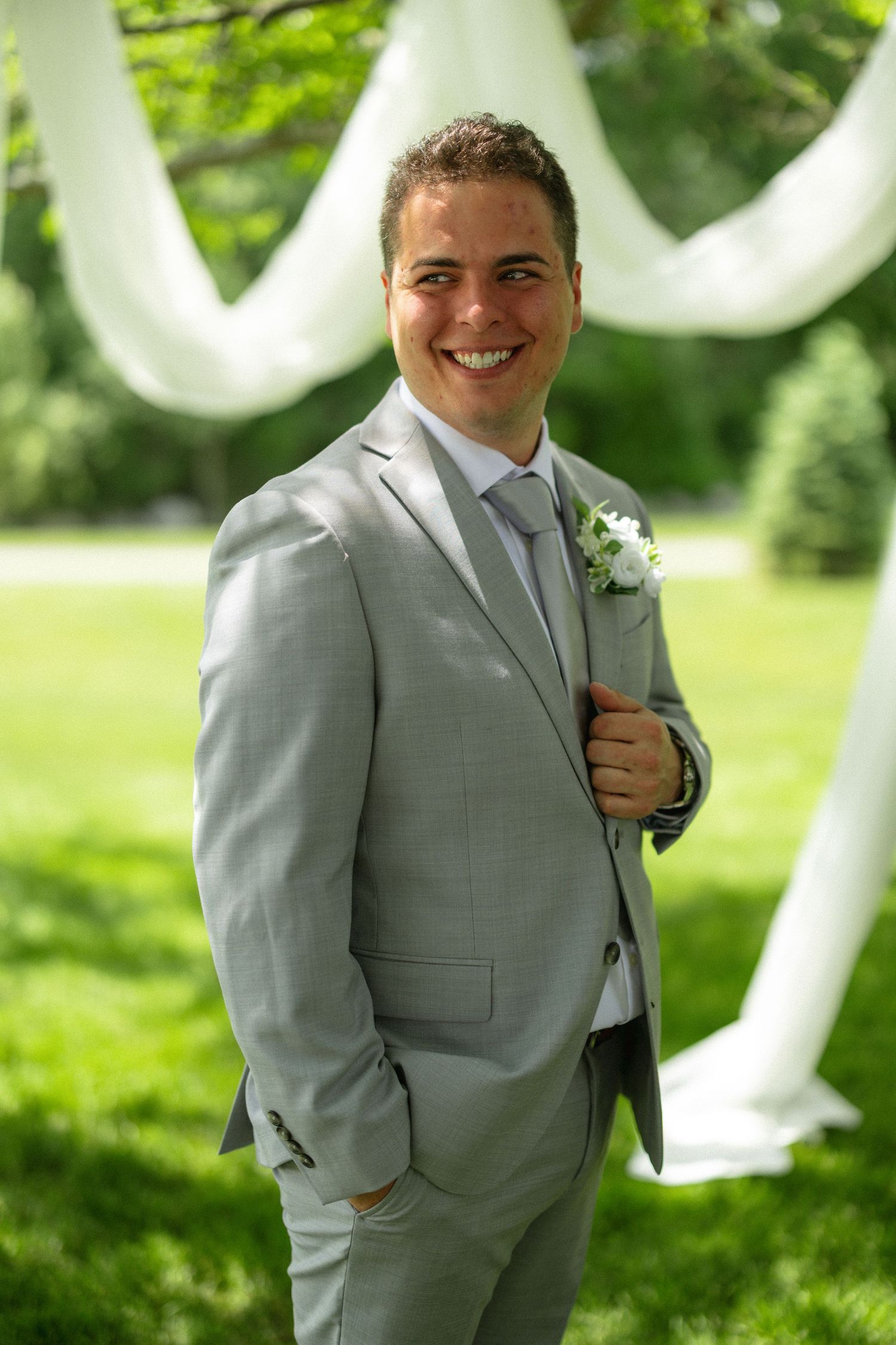 Smiling groom in light gray suit with boutonniere posing outdoors before wedding ceremony.