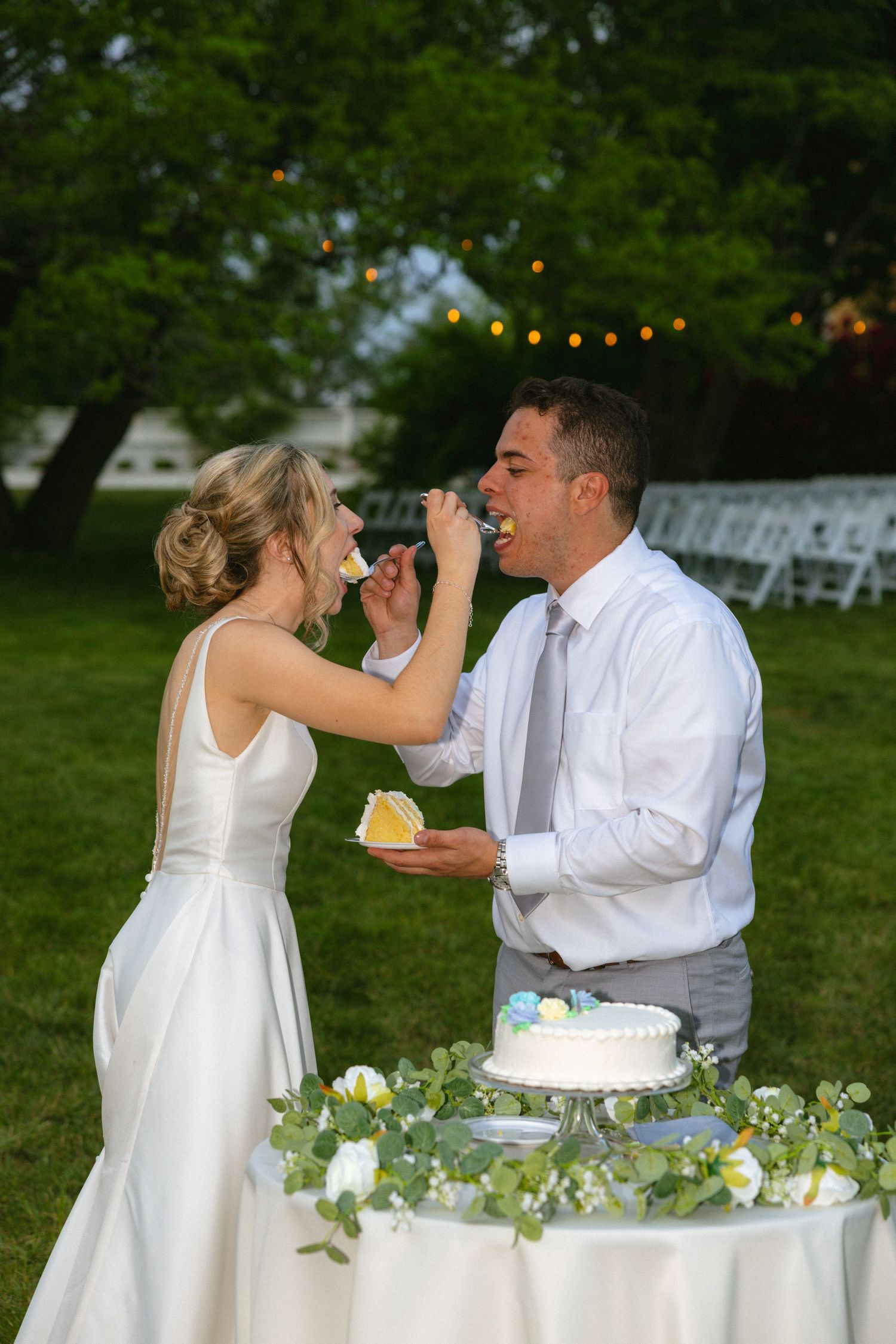 Bride feeds groom wedding cake slice at outdoor reception with white chairs and string lights.