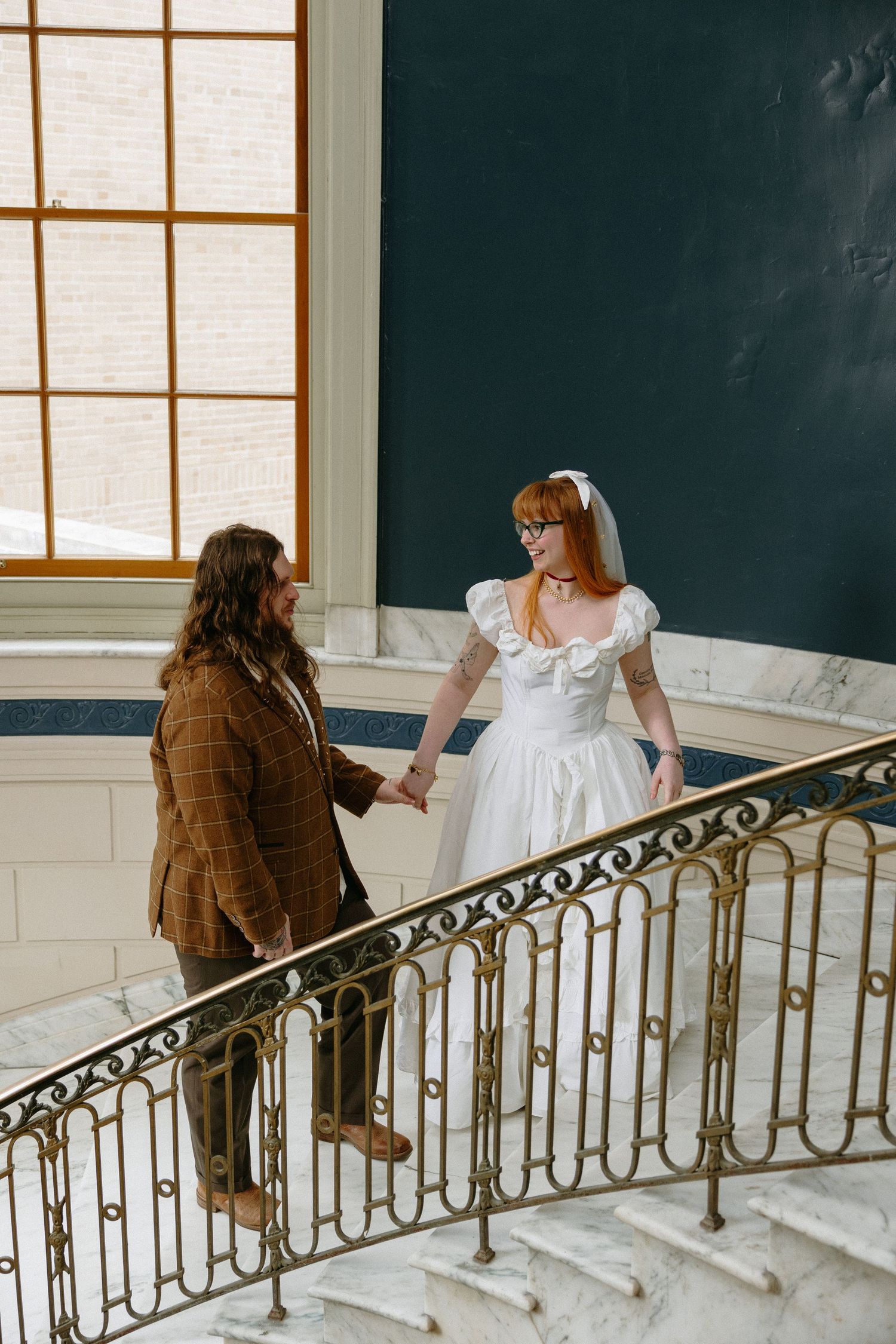 A redheaded bride in a white gown holds hands with a groom in a brown suit on an elegant marble staircase.