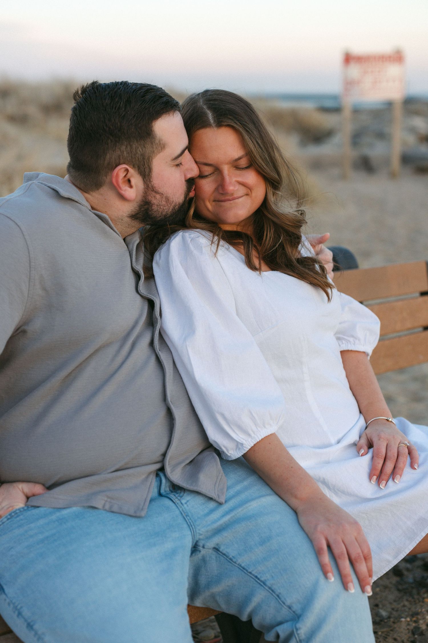 Couple sitting on a bench at the beach, man kissing woman's cheek as she smiles with eyes closed.