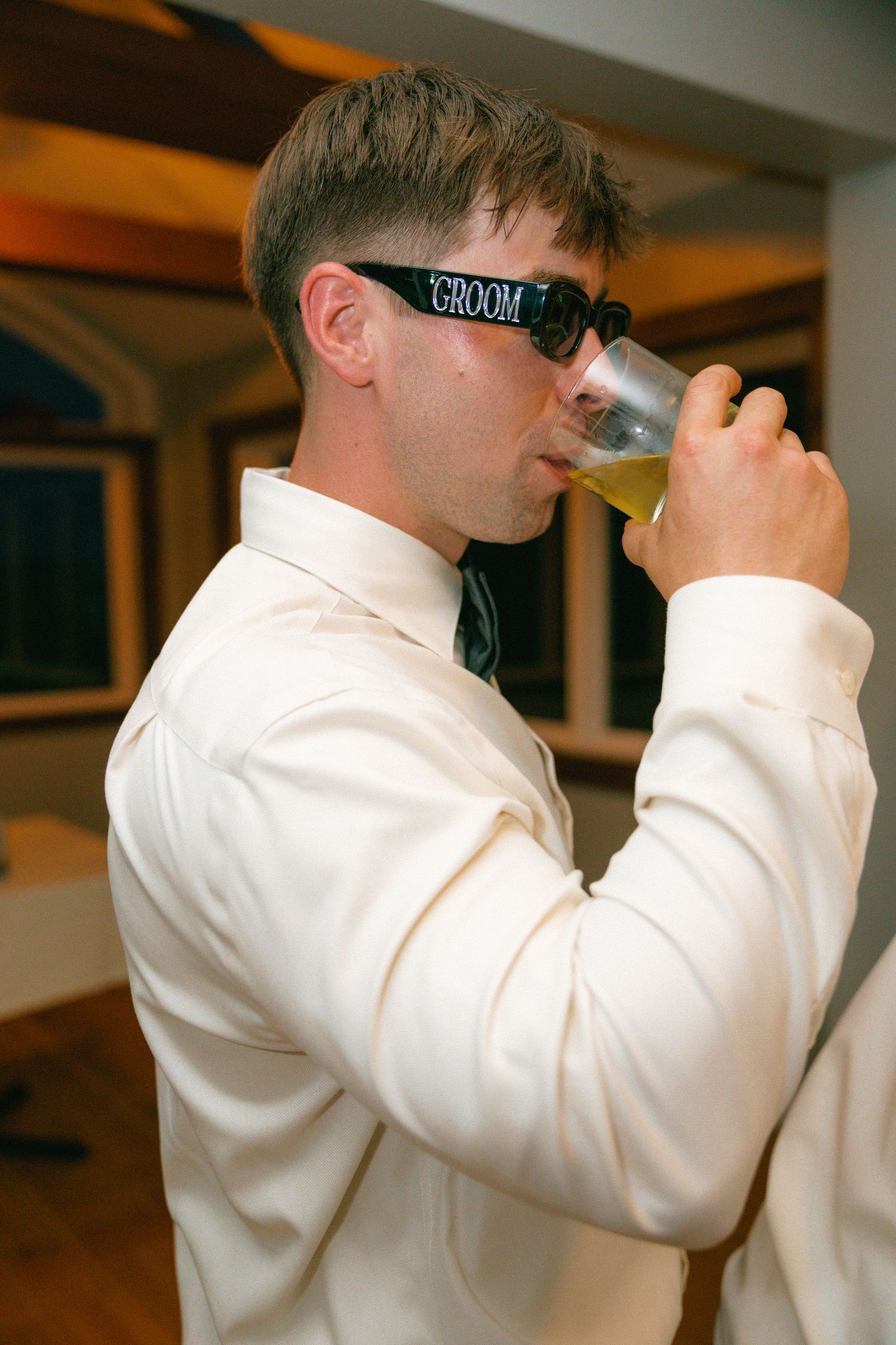 Groom wearing novelty sunglasses drinks beer in white shirt at wedding celebration.
