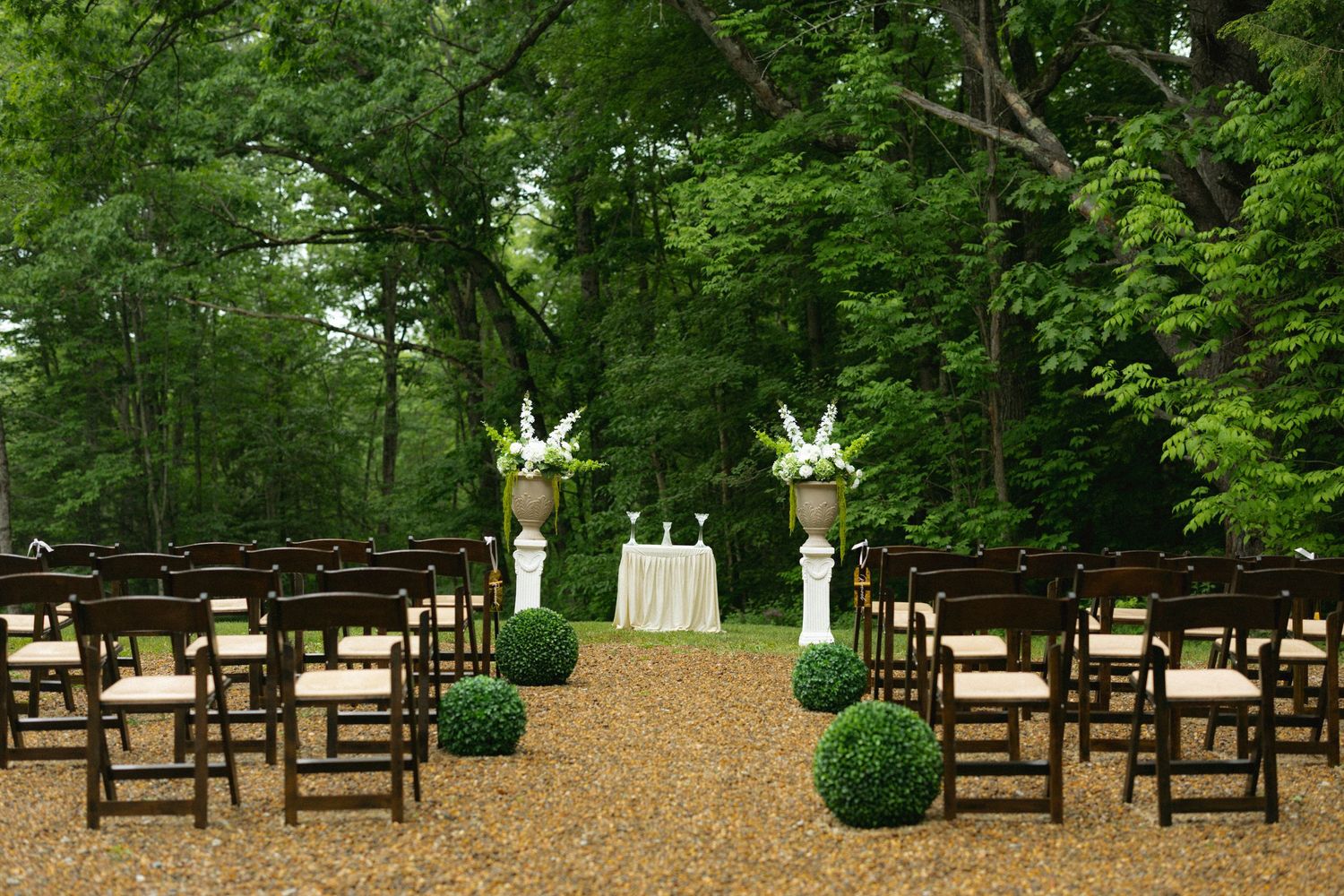 Elegant outdoor wedding ceremony setup with dark wood chairs, gravel aisle, boxwood topiaries, and floral arrangements in wooded setting.
