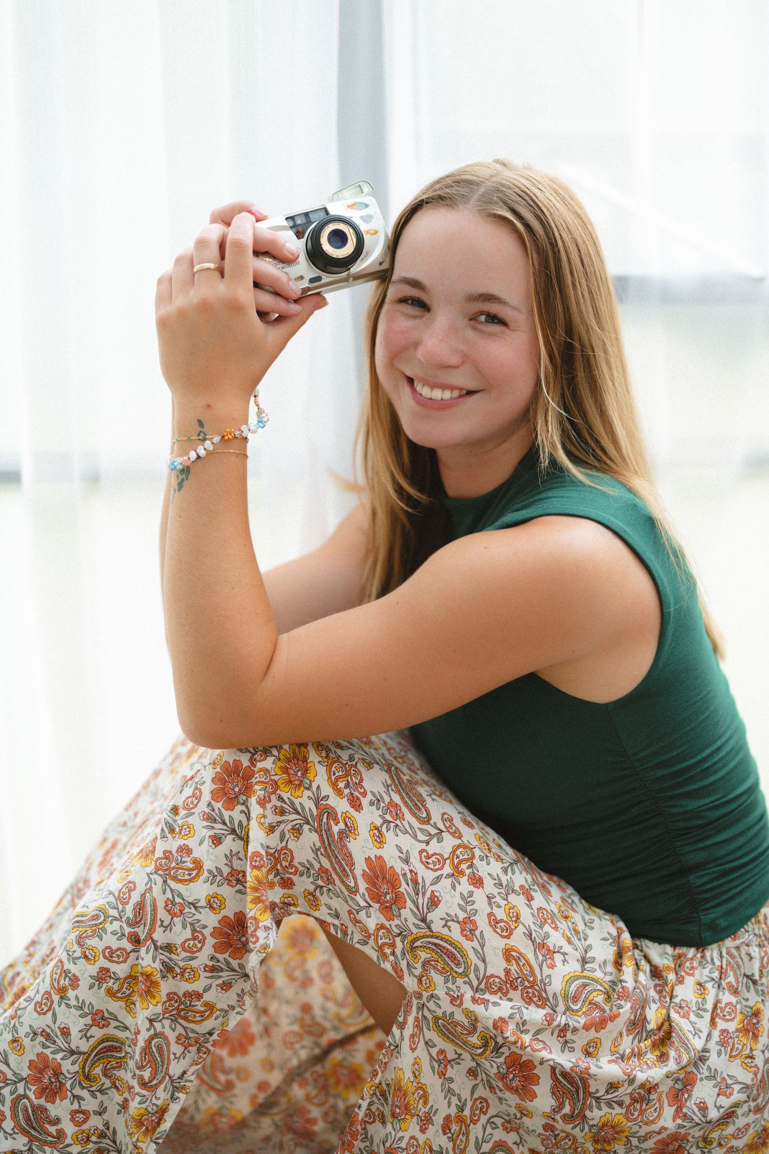 Smiling teenage girl holding a film camera, wearing a green top and floral skirt, sitting near a bright window.