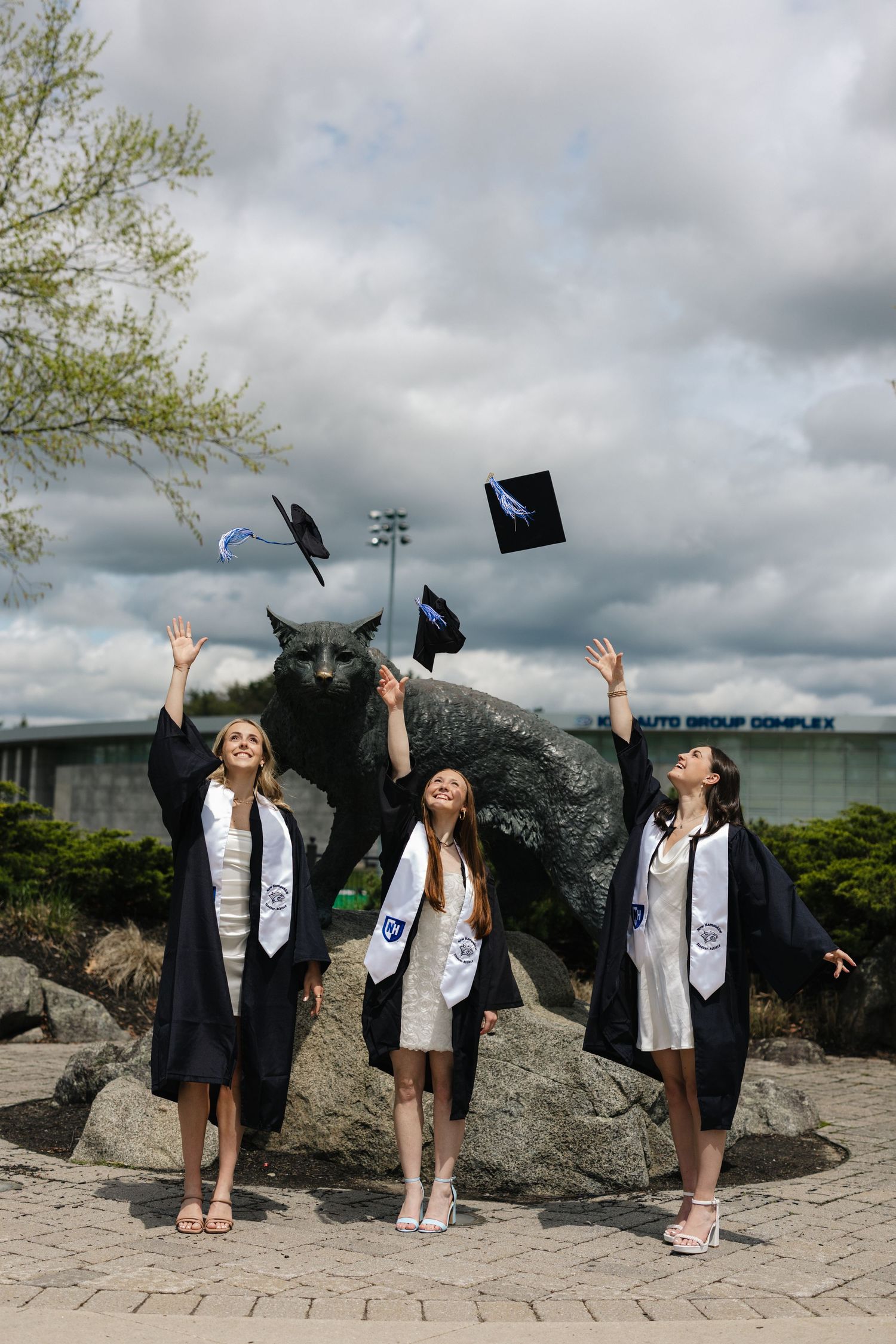 Graduates throwing caps in the air near large stone sculpture on cloudy day.