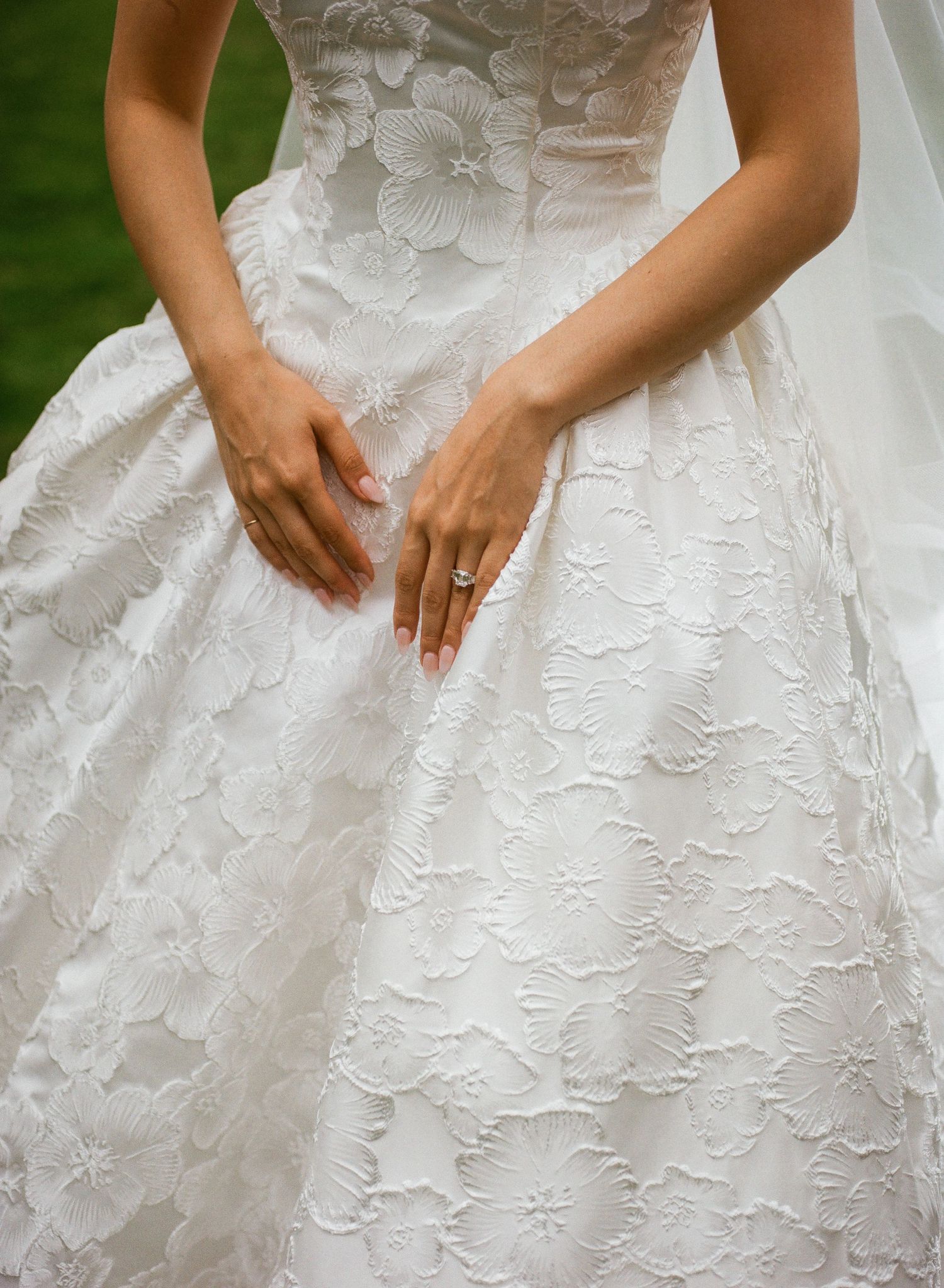 Bride in a white floral lace ball gown wedding dress, hands resting on skirt, wearing a diamond engagement ring.