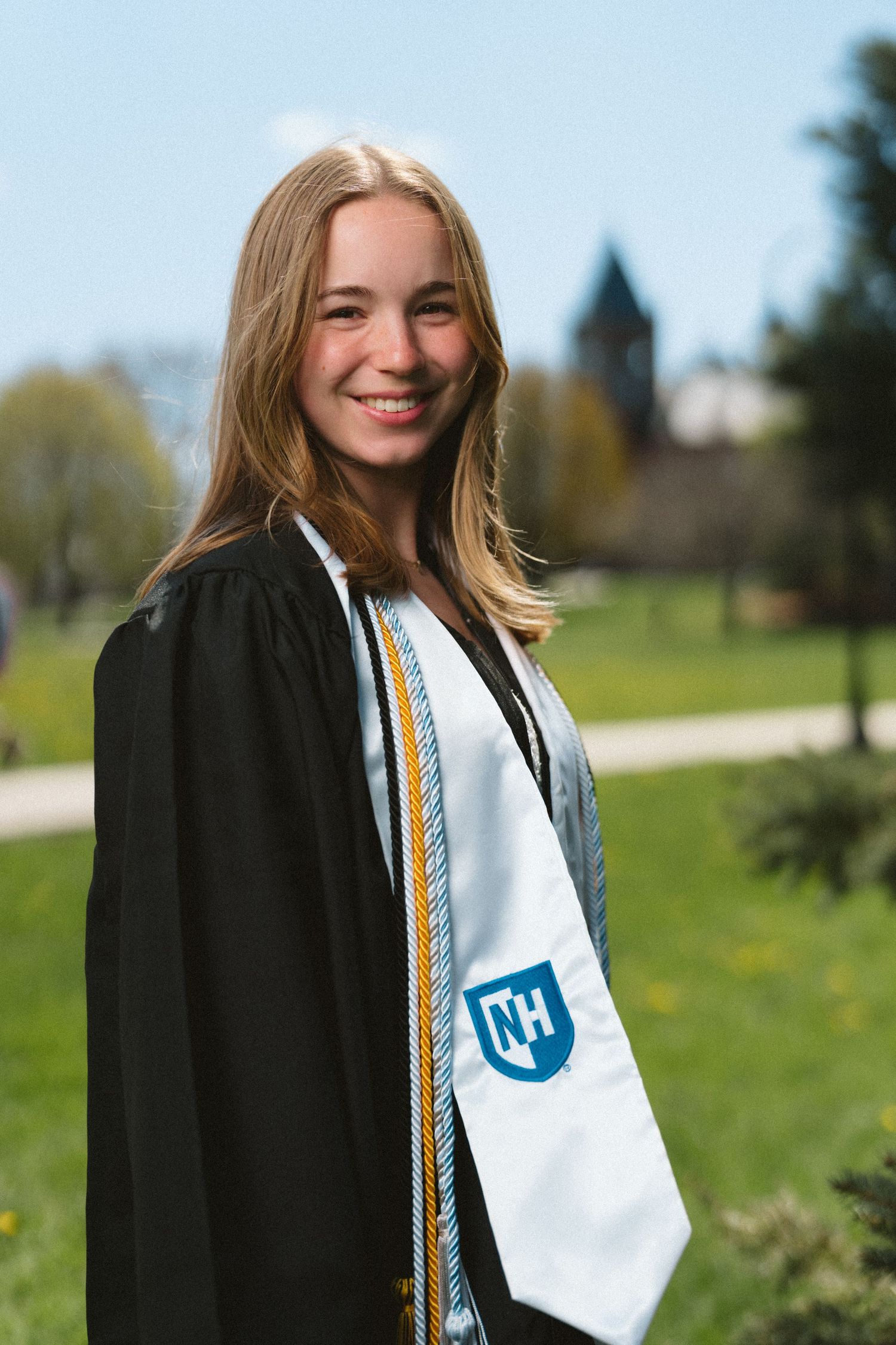Smiling graduate in black cap and gown with white stole outdoors on a sunny day.