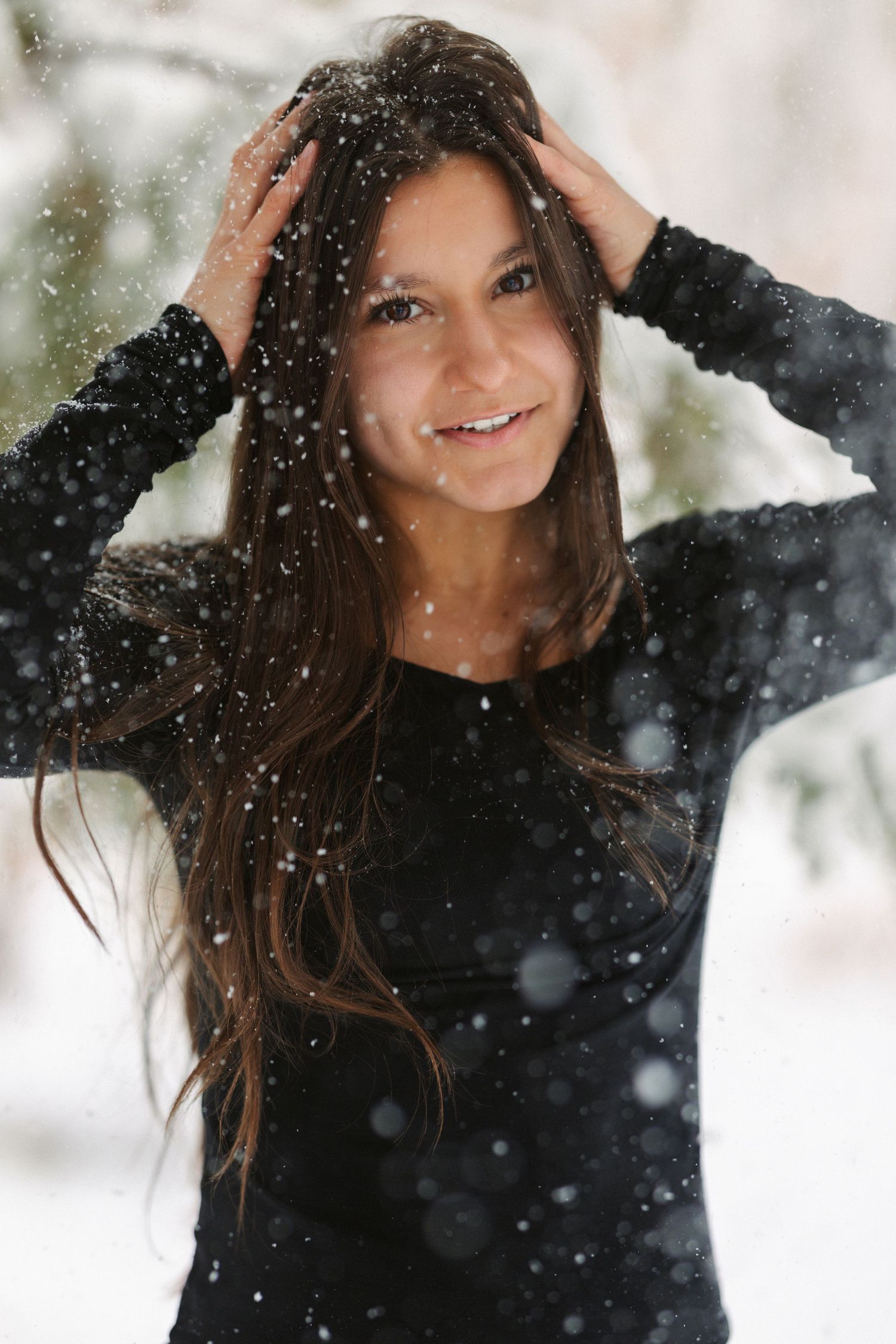 A person in a black sweater smiles and plays with their long dark hair while snowflakes fall in a winter scene.