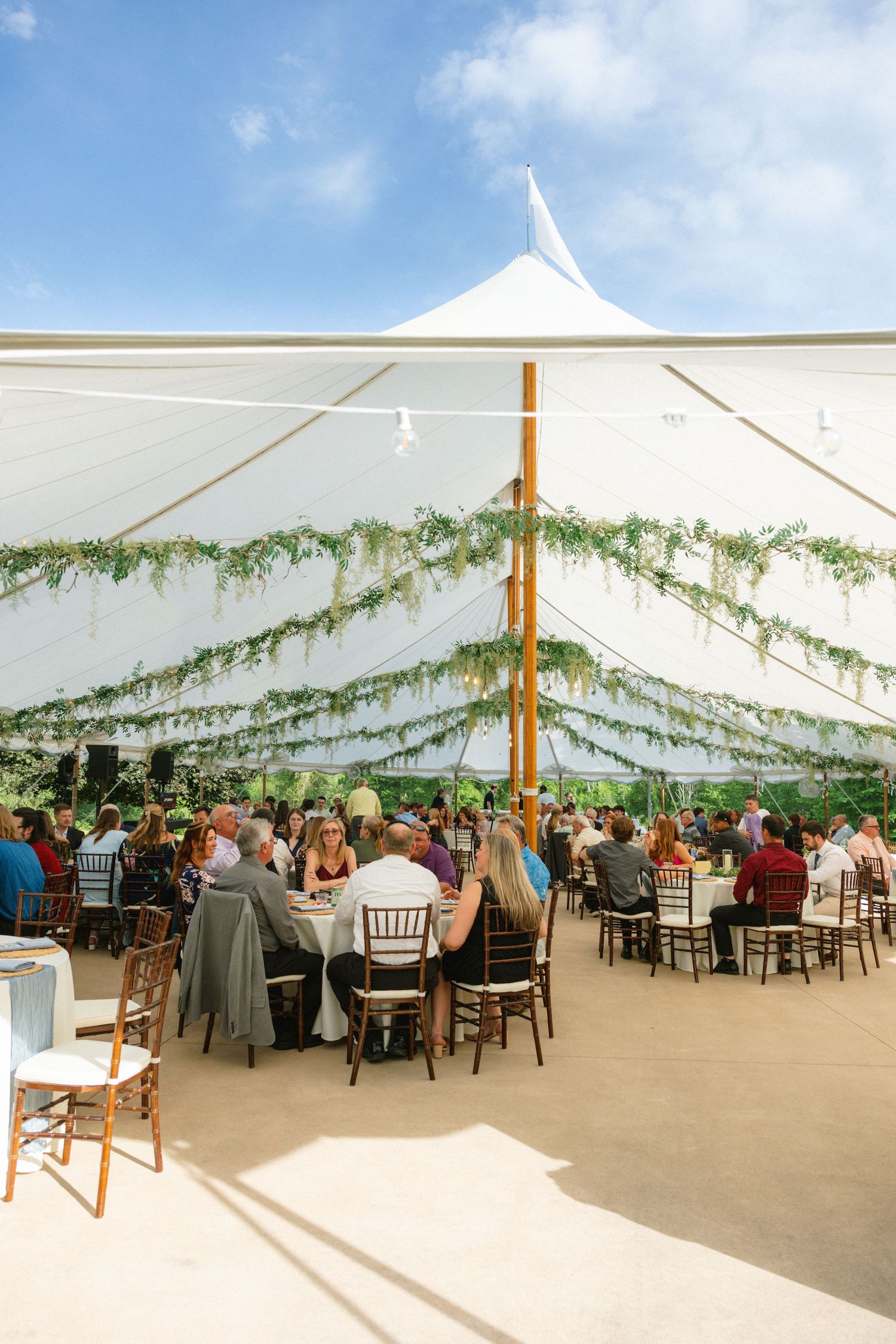 Elegant outdoor wedding reception under a white tent with greenery garlands and guests dining.