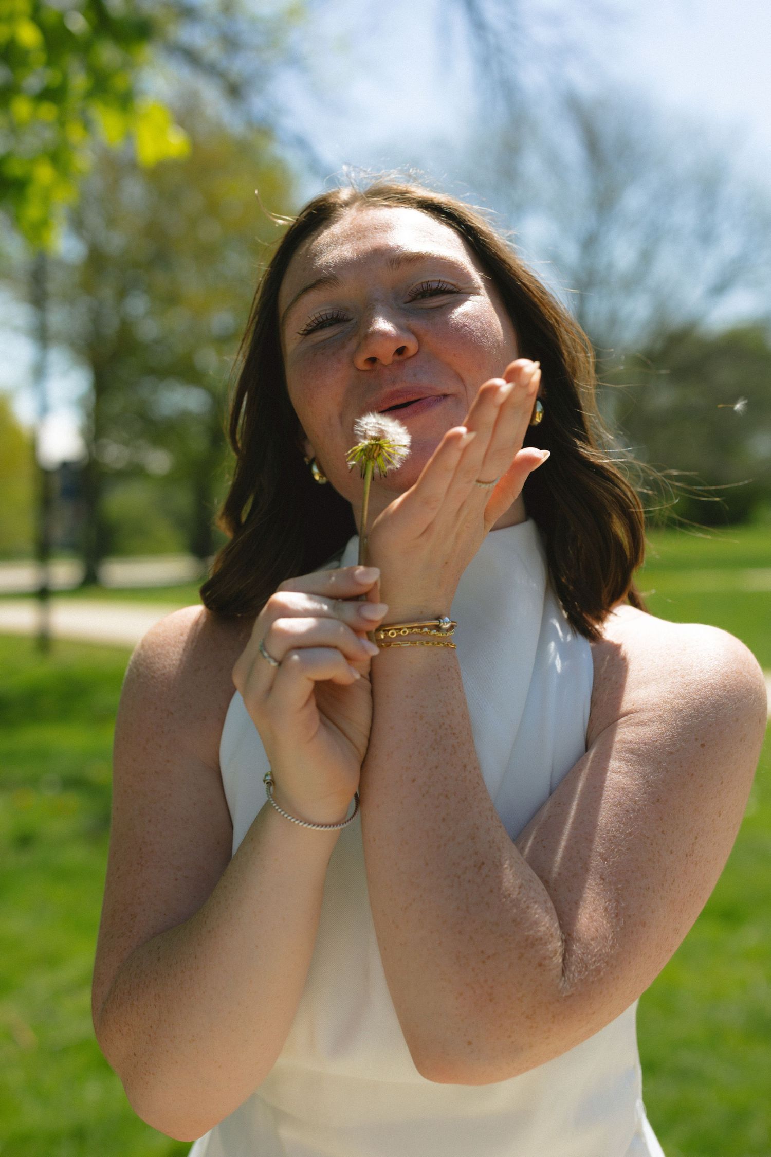 Young woman in white dress blowing dandelion seeds in a sunny green park, smiling with eyes closed.