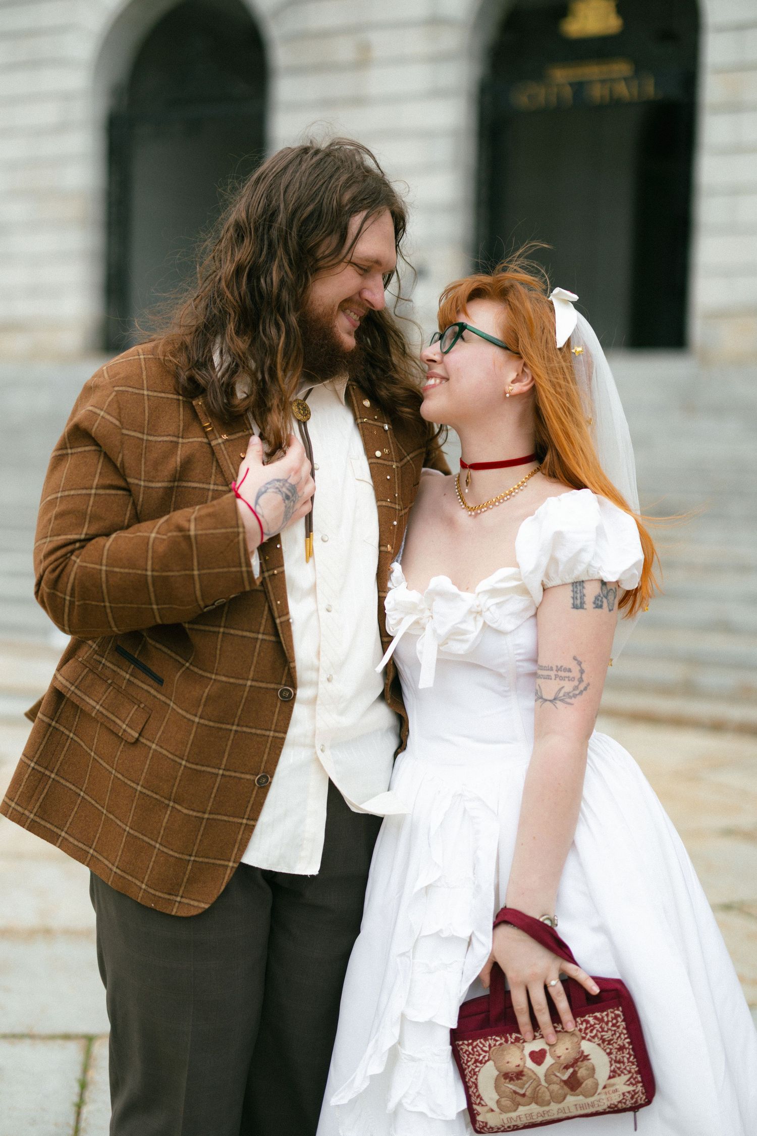A bride in white dress and groom in brown plaid jacket share a romantic moment outside a city hall building.