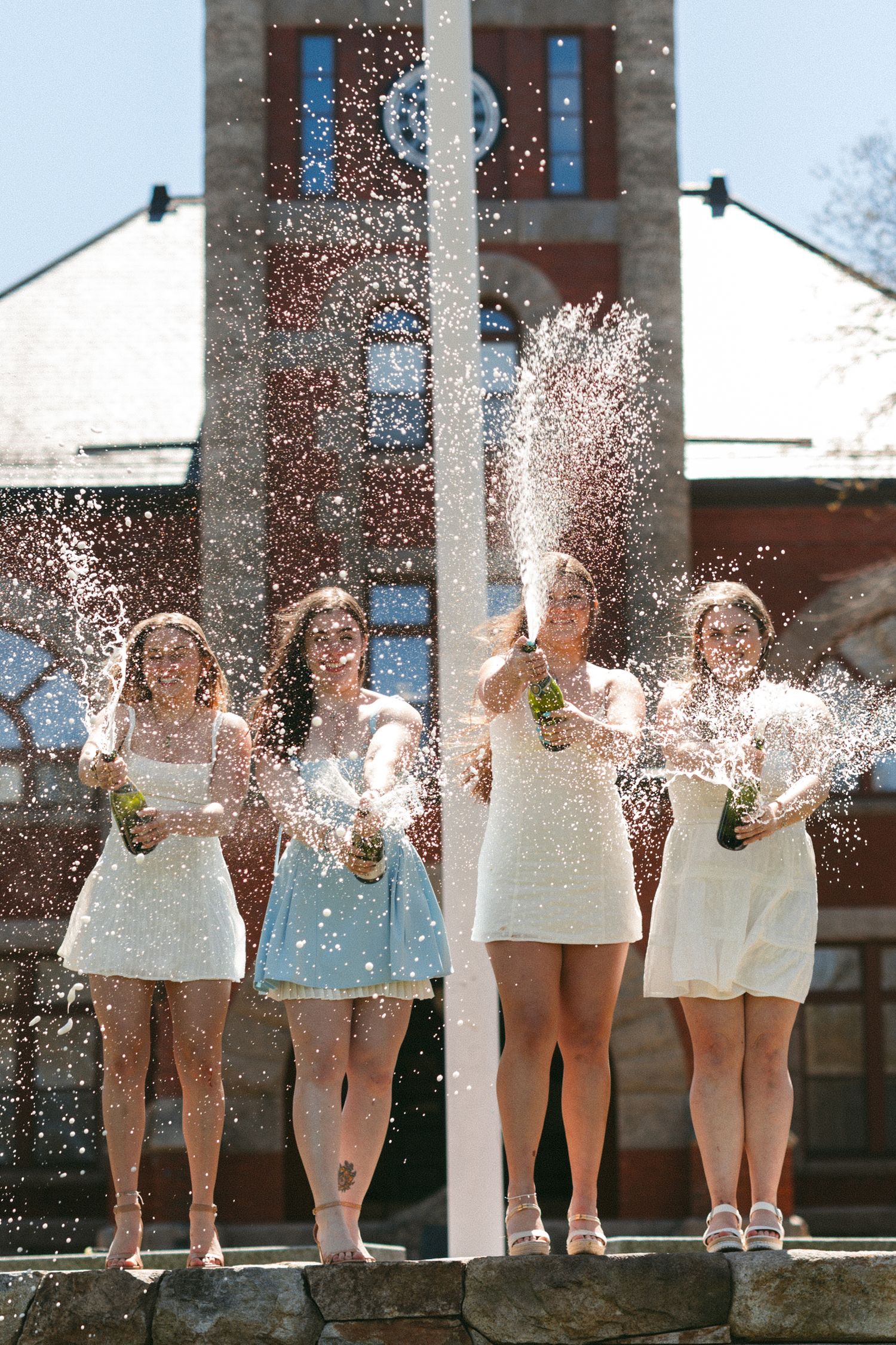 Four young women in white dresses celebrate outdoors by spraying champagne bottles in front of a brick building.