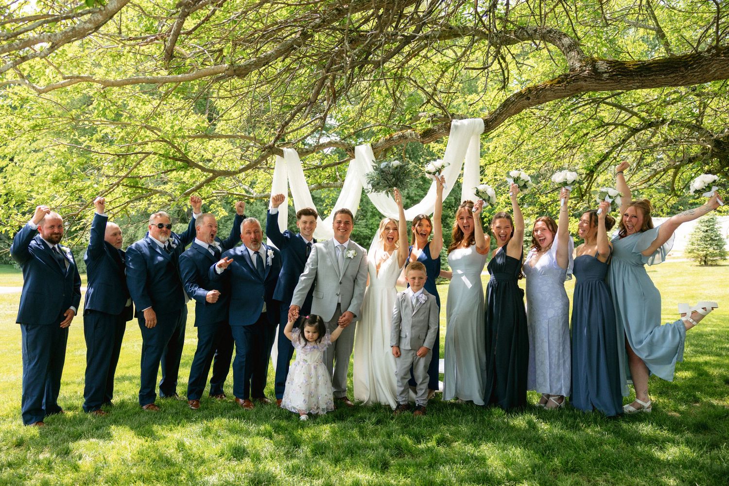 Large wedding party celebrating under a sprawling tree, tossing bouquets and cheering in blue and gray attire.