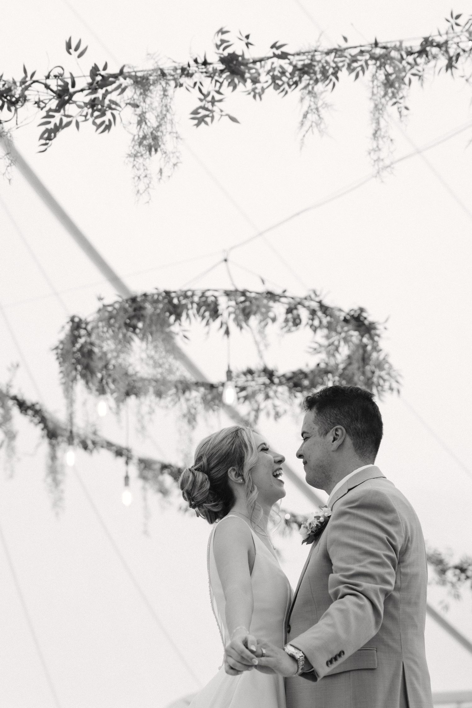 Black and white photo of smiling bride and groom sharing first dance under floral chandelier in elegant tent.
