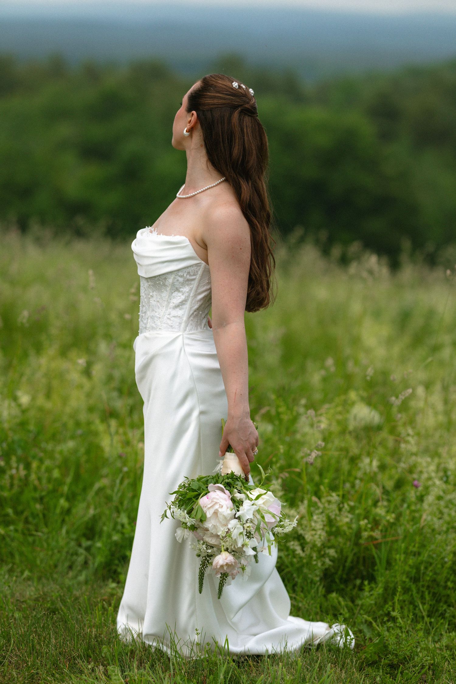 Bride in strapless white gown holds bouquet while standing in a meadow with scenic green hills behind her.
