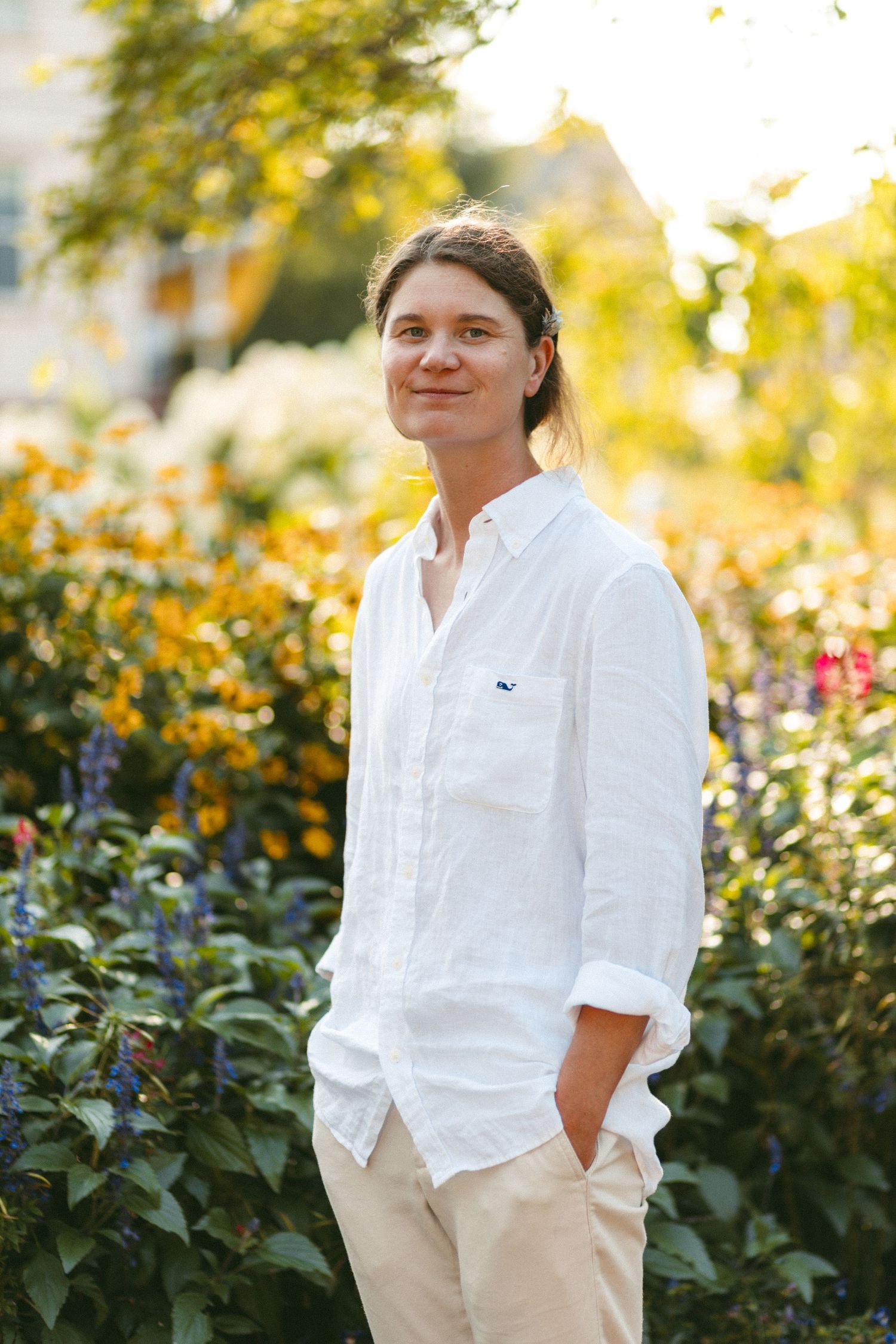 Bride in white shirt and khaki pants poses in front of blooming garden.