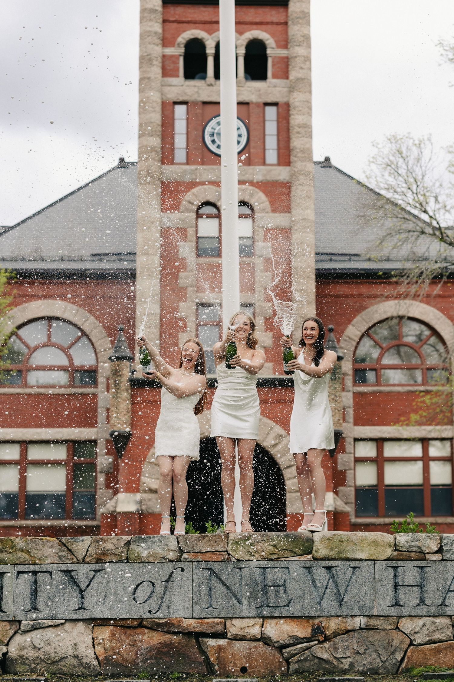 Graduates celebrating on stone wall with water splashing in front of brick building.