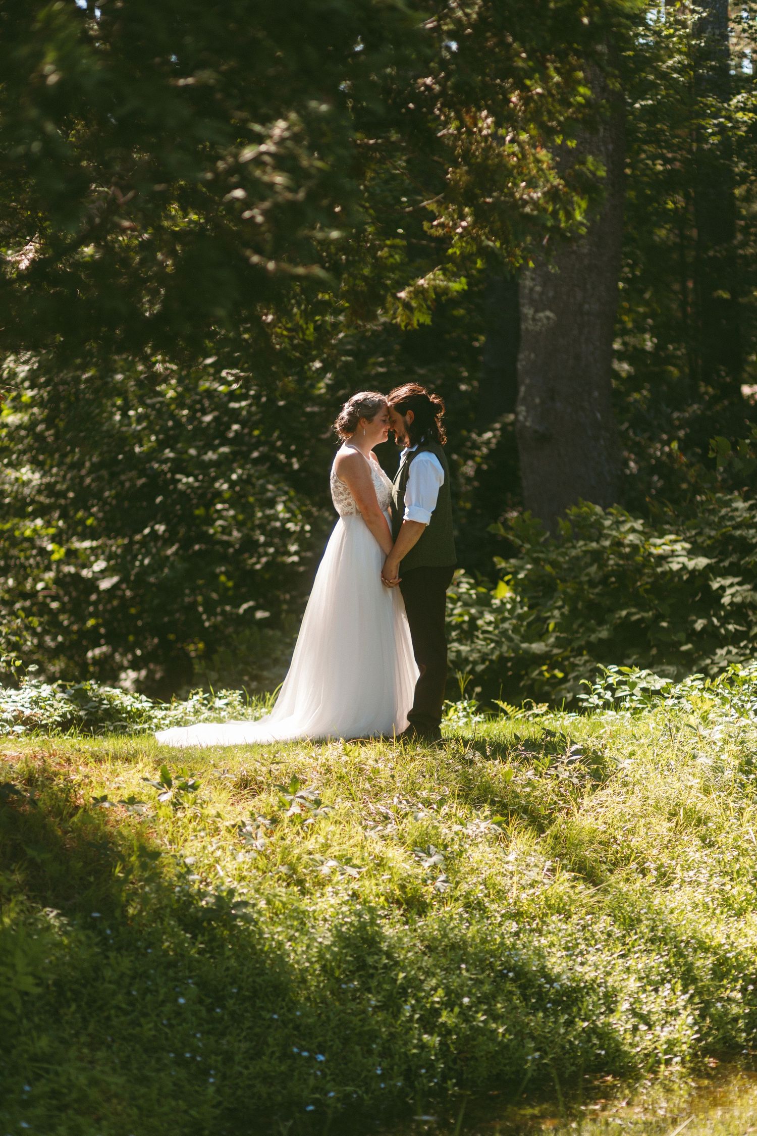 A couple in wedding attire stands together in a sunlit garden setting with lush greenery.