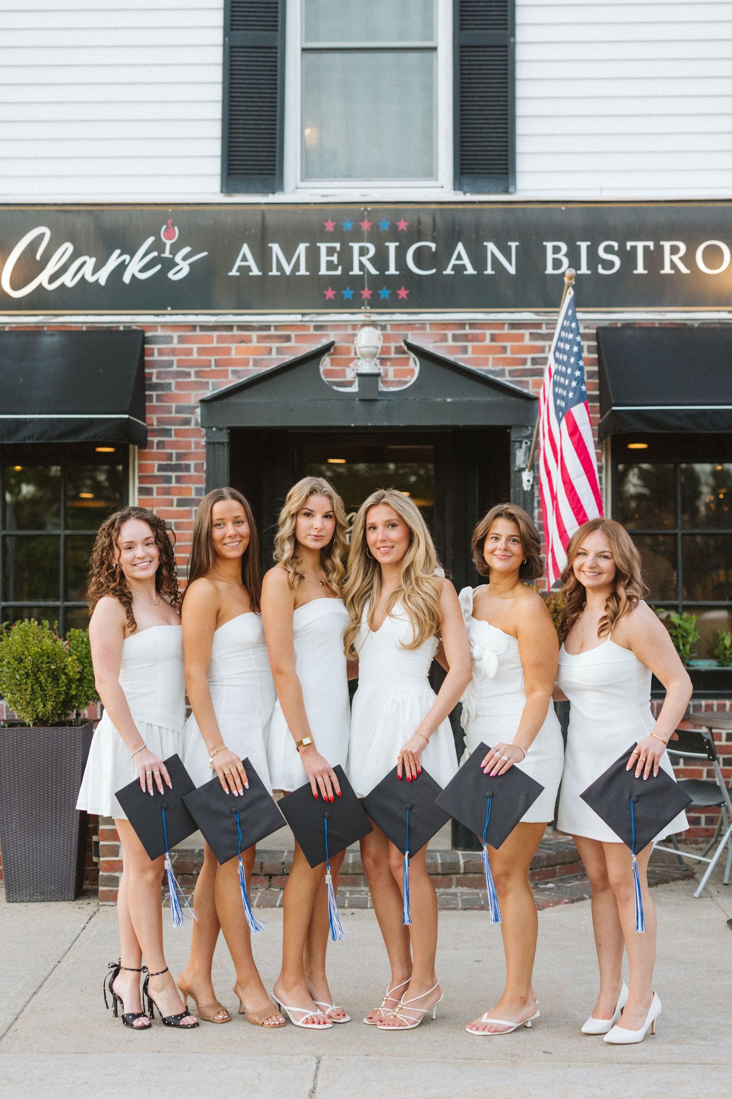 Six young women in white dresses holding graduation caps pose in front of Clark's American Bistro with an American flag.
