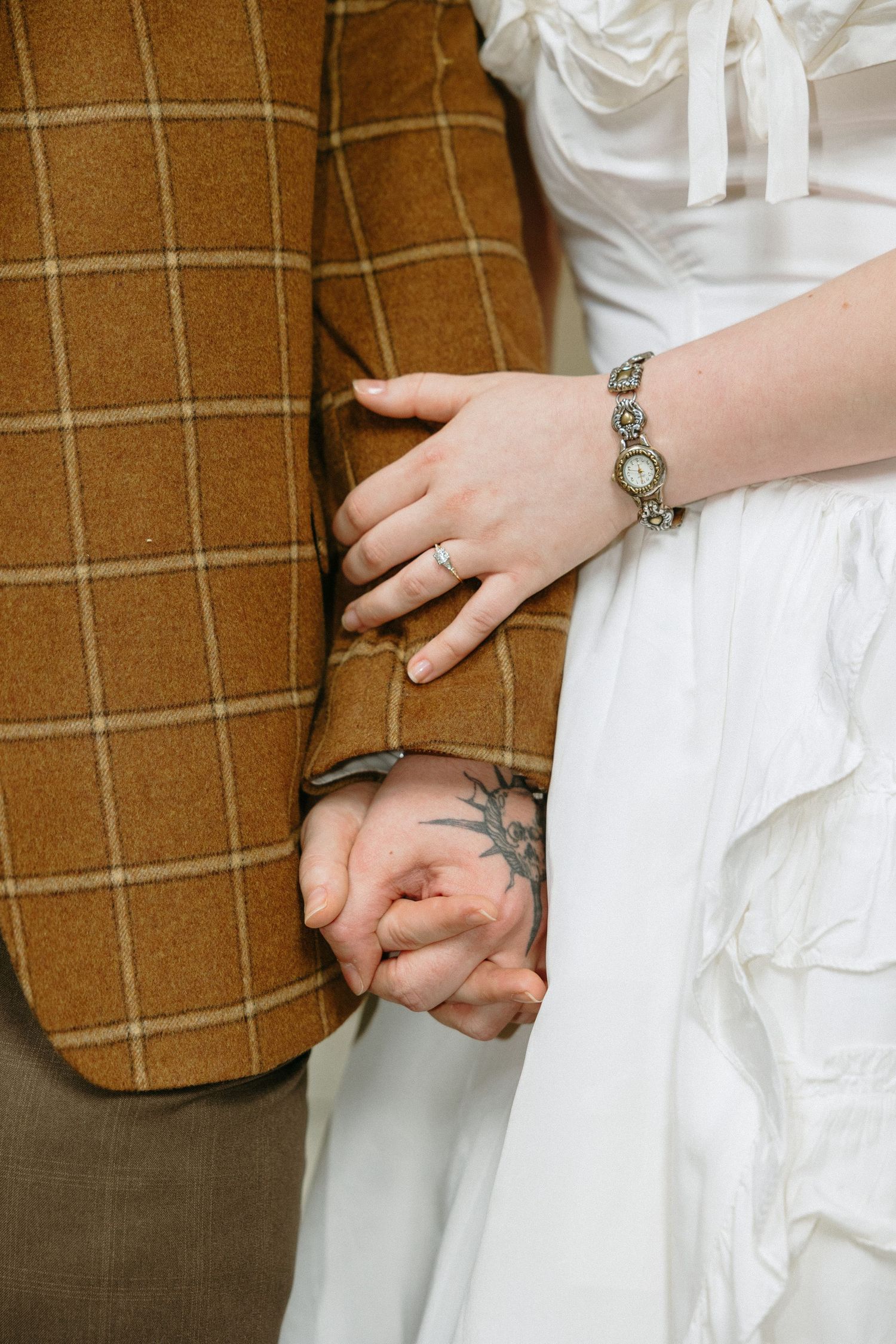 Couple holding hands at wedding, bride in white dress with silver watch and ring, groom in brown plaid blazer, tattoo visible.