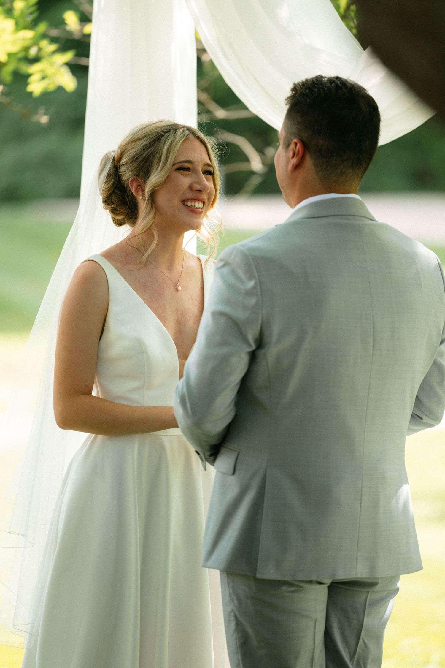 Smiling bride in white V-neck gown holds hands with groom in gray suit during outdoor wedding ceremony.