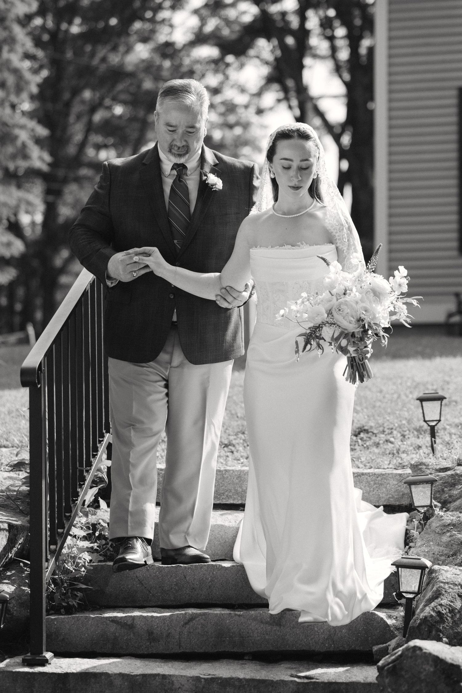 Black and white photo of father escorting bride in white gown and veil down stone steps holding bouquet.