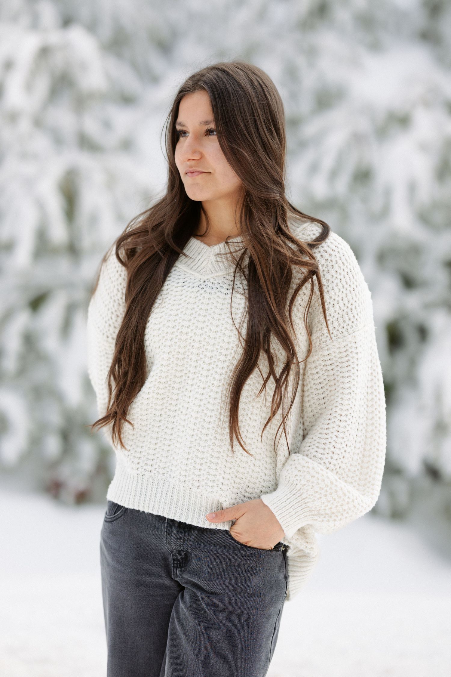 Person wearing a cozy white knit sweater poses against a snowy winter background with evergreen trees.