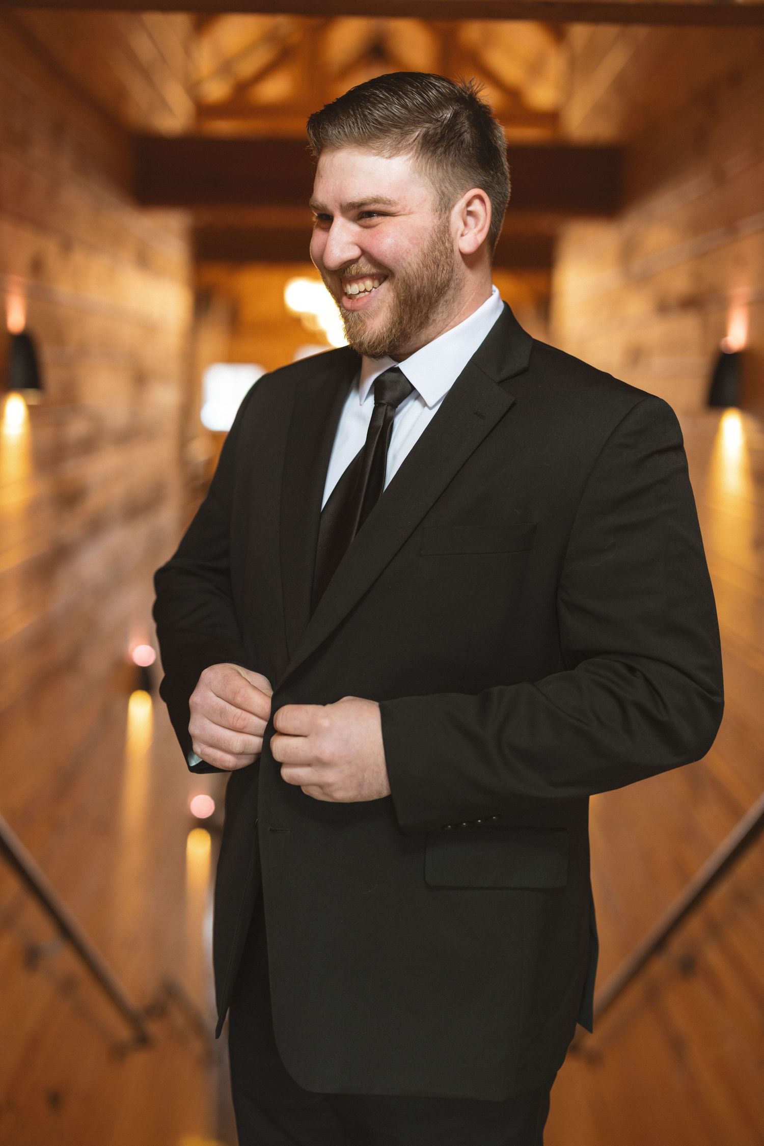 Person in black suit adjusts their jacket buttons in warm-lit indoor setting with wooden background.