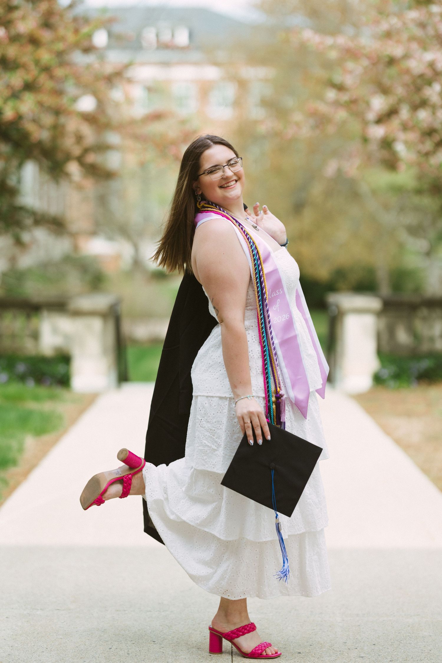 Smiling graduate in white dress holds cap, wearing black gown and colorful stoles, posing outdoors on campus path.