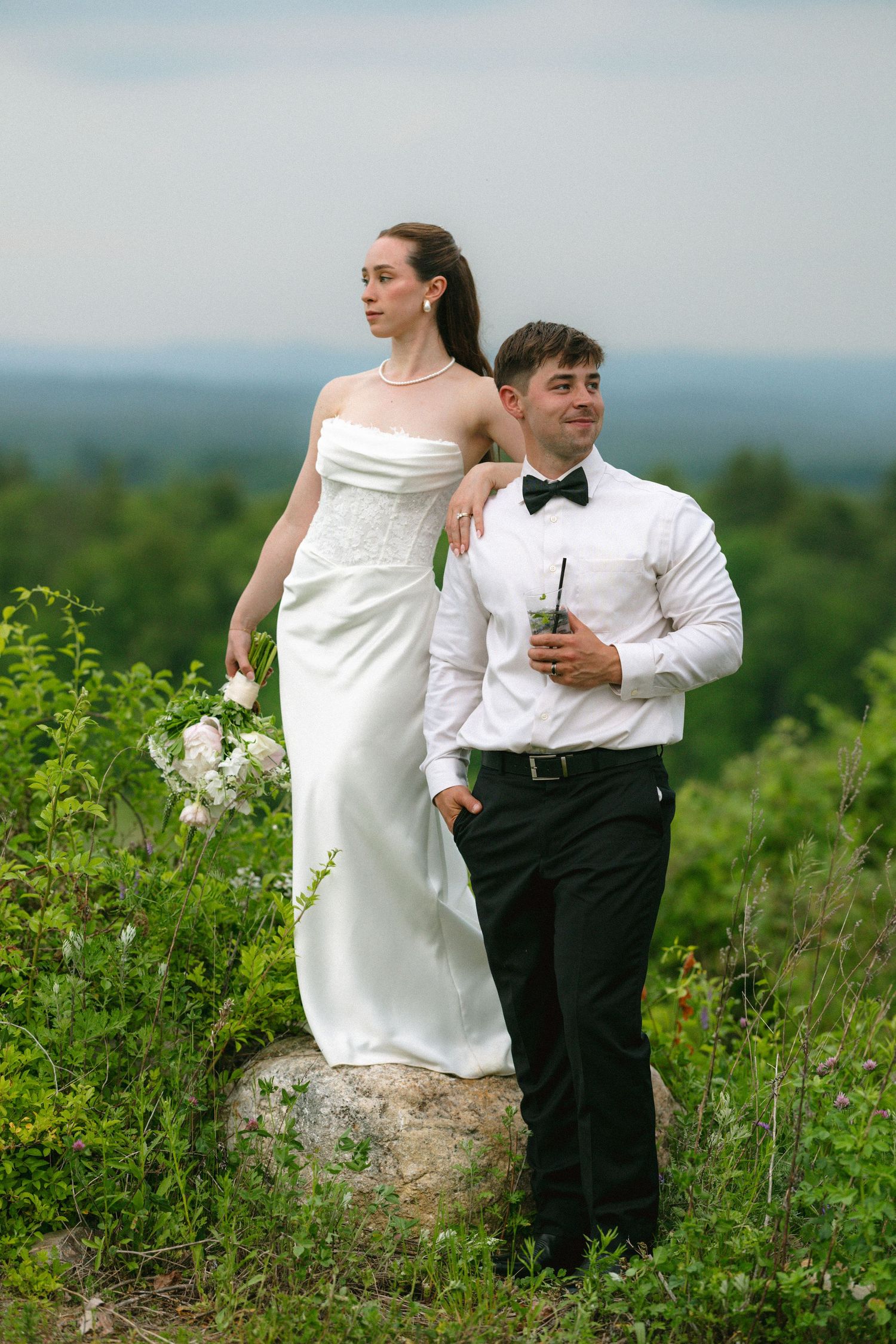 Bride in white gown stands on rock with groom in tuxedo against lush green landscape backdrop.