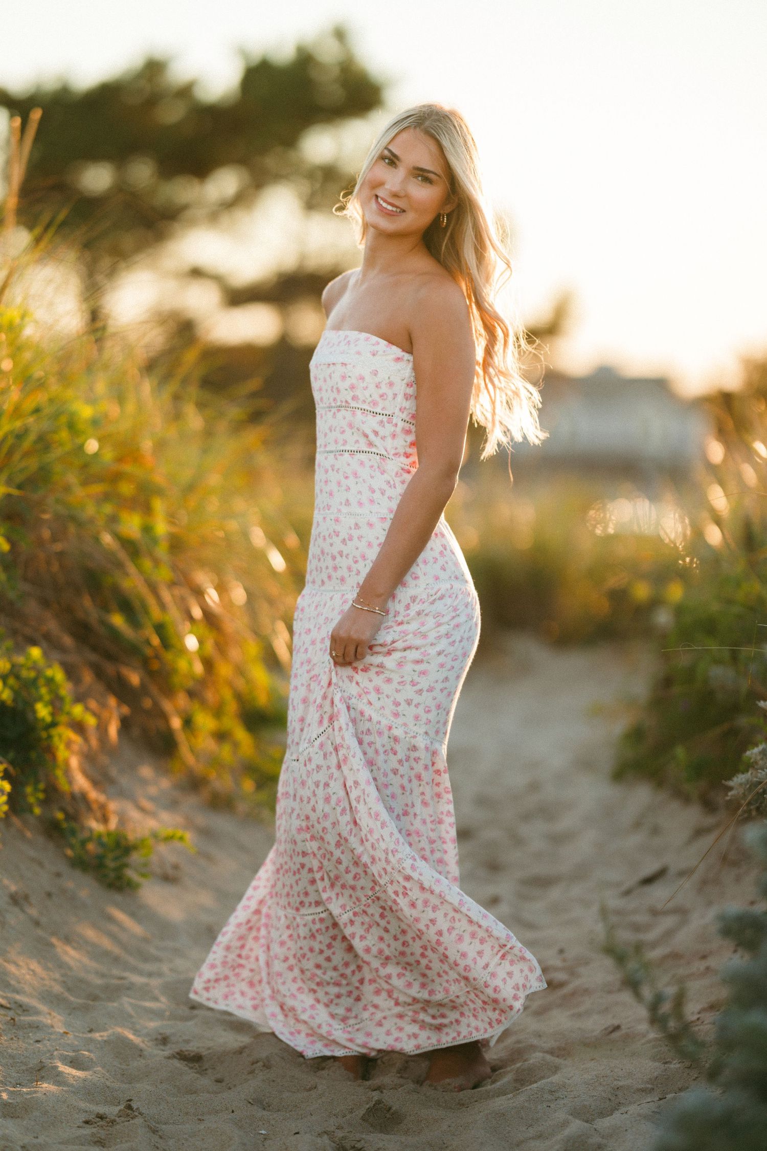 Sunset beach portraits featuring someone in a flowing white floral dress among coastal vegetation.