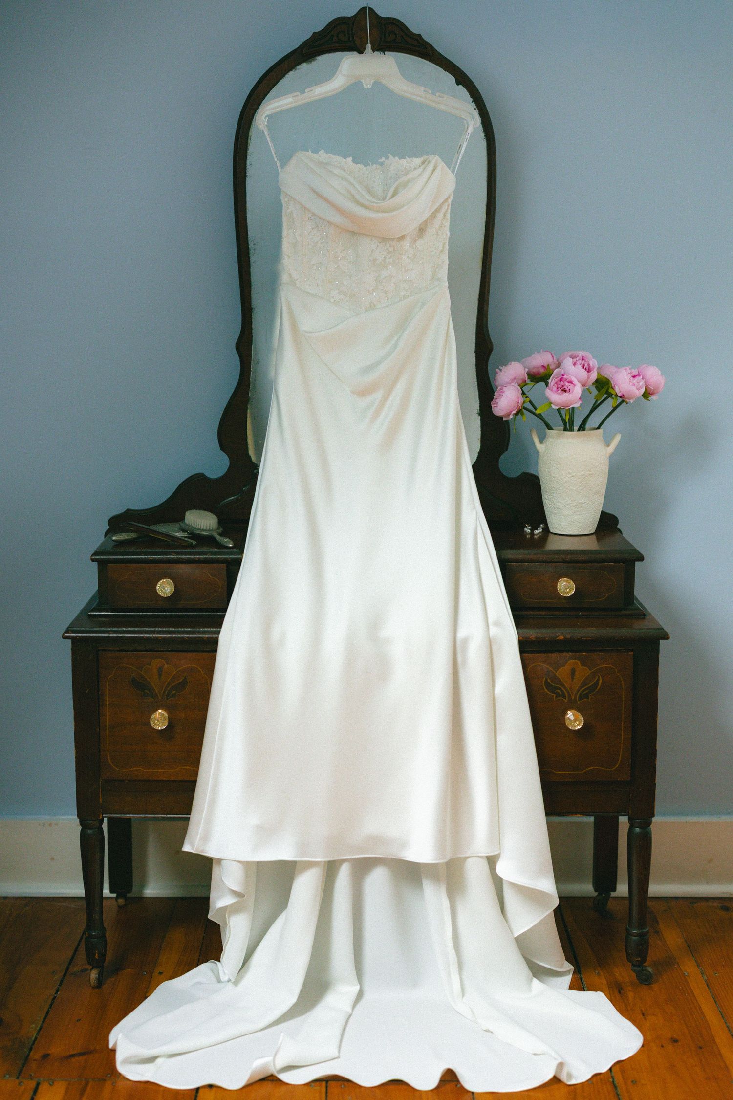 Elegant strapless wedding dress hanging on antique dark wood dresser mirror with pink peonies in white vase.