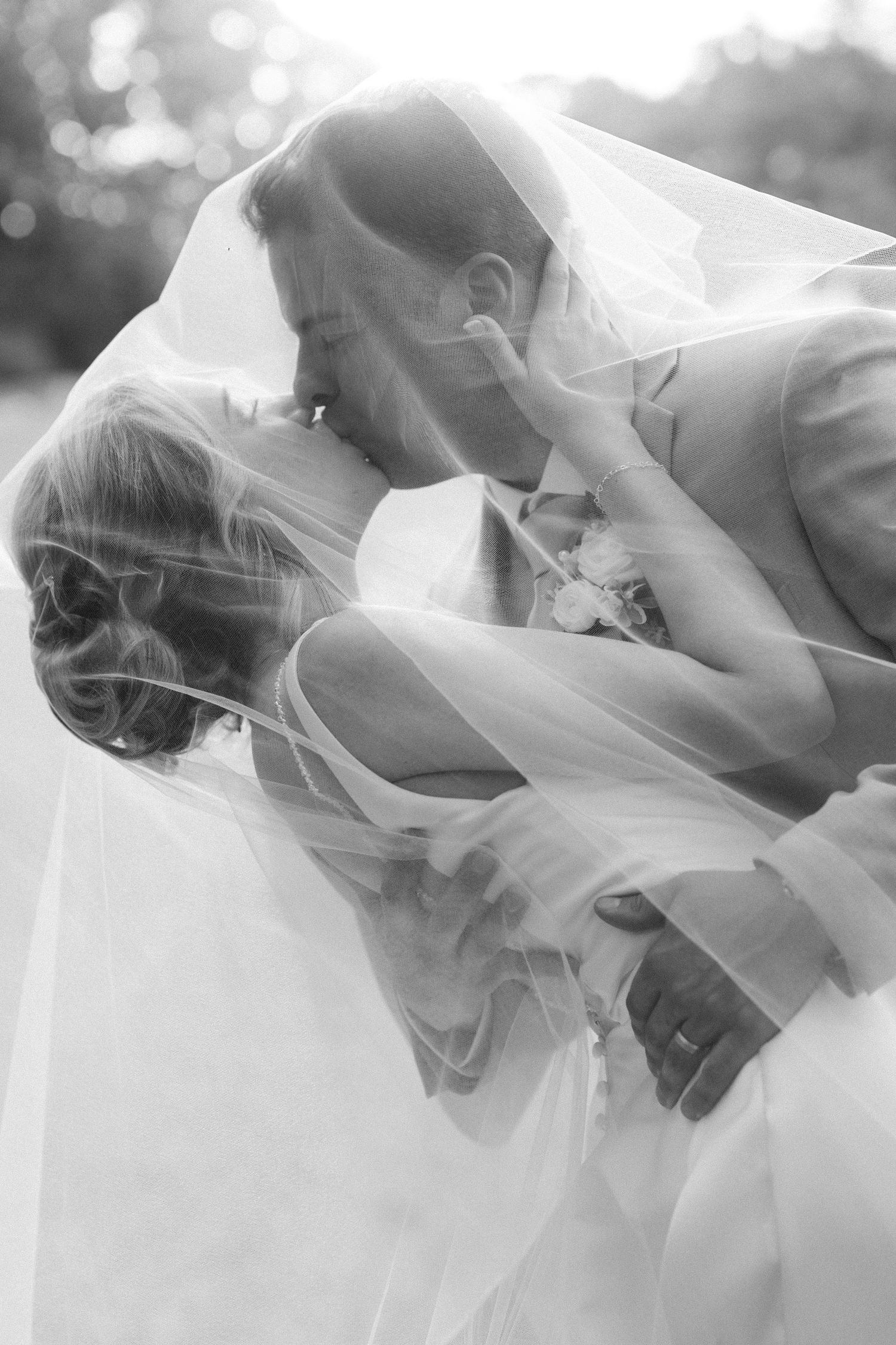 Black and white photo of bride and groom sharing a romantic kiss under flowing wedding veil outdoors.
