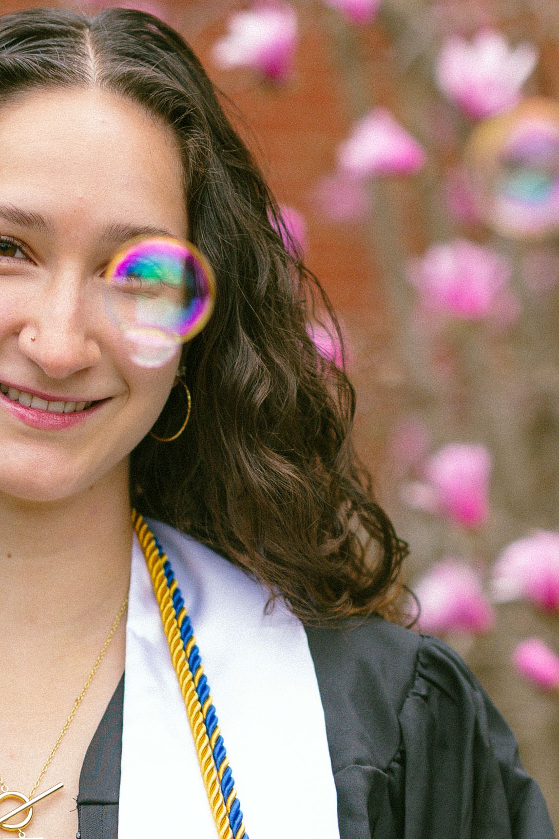 Smiling graduate in black gown with gold and blue cords poses with a soap bubble near pink magnolia blossoms.