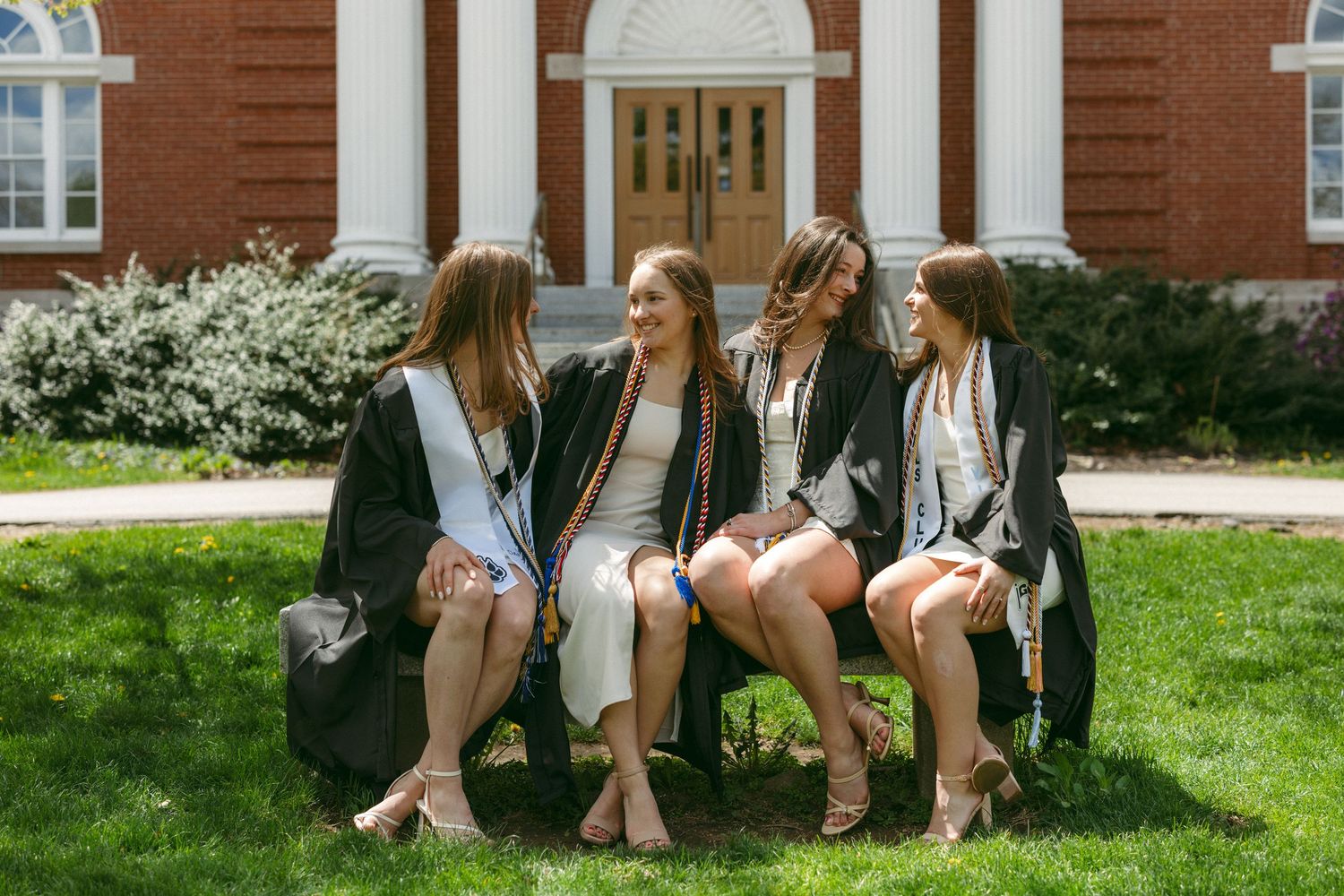 Four female graduates in black gowns sit on a bench outside a brick building, smiling and chatting together.