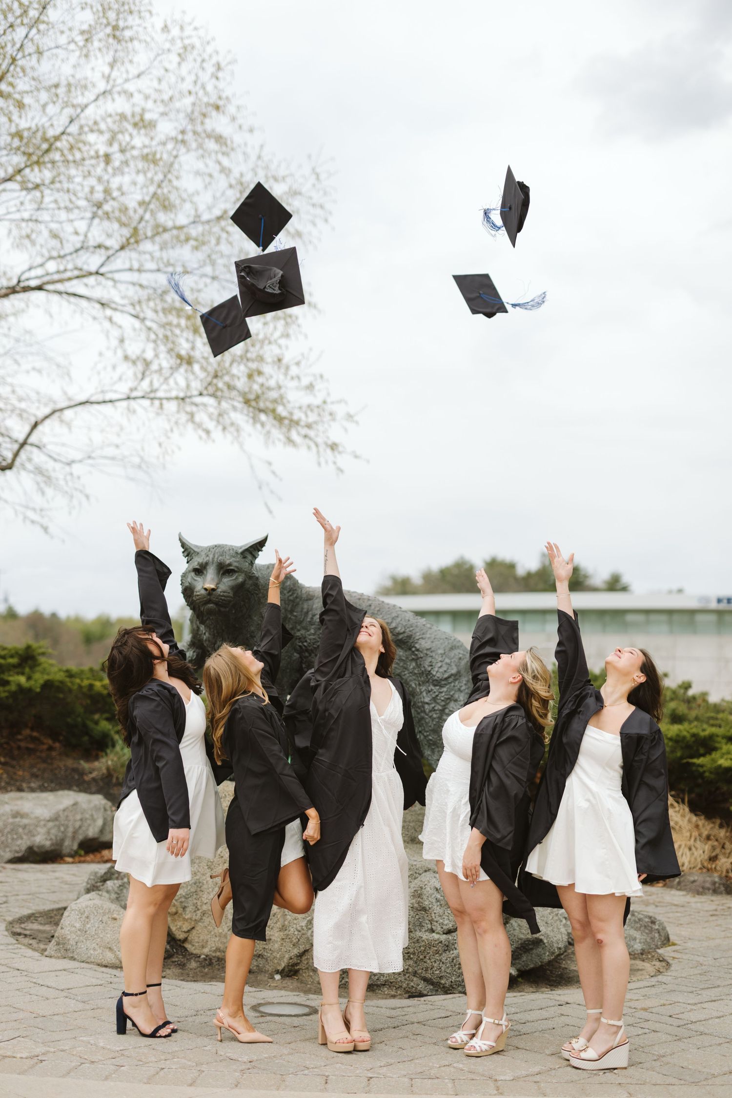 Five graduates in white dresses toss black mortarboard caps in the air near a wildcat statue outdoors.
