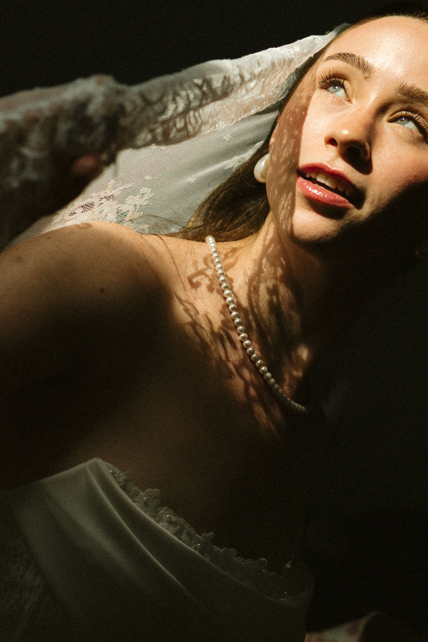 A woman in a white lace veil and pearl necklace gazes upward dramatically in moody, shadowed lighting.
