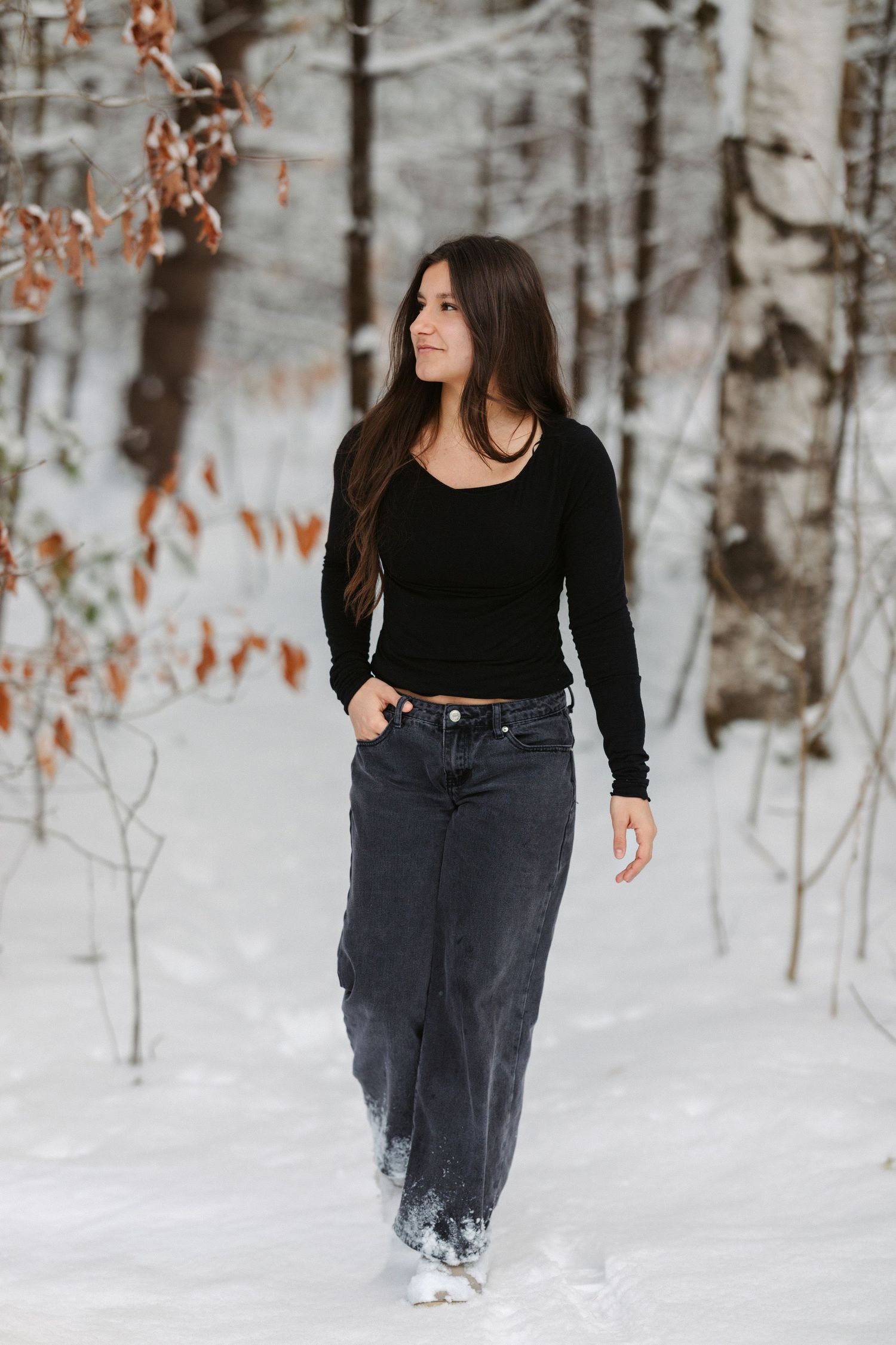 Person in black winter outfit poses in snowy forest with bare trees and autumn leaves in background.