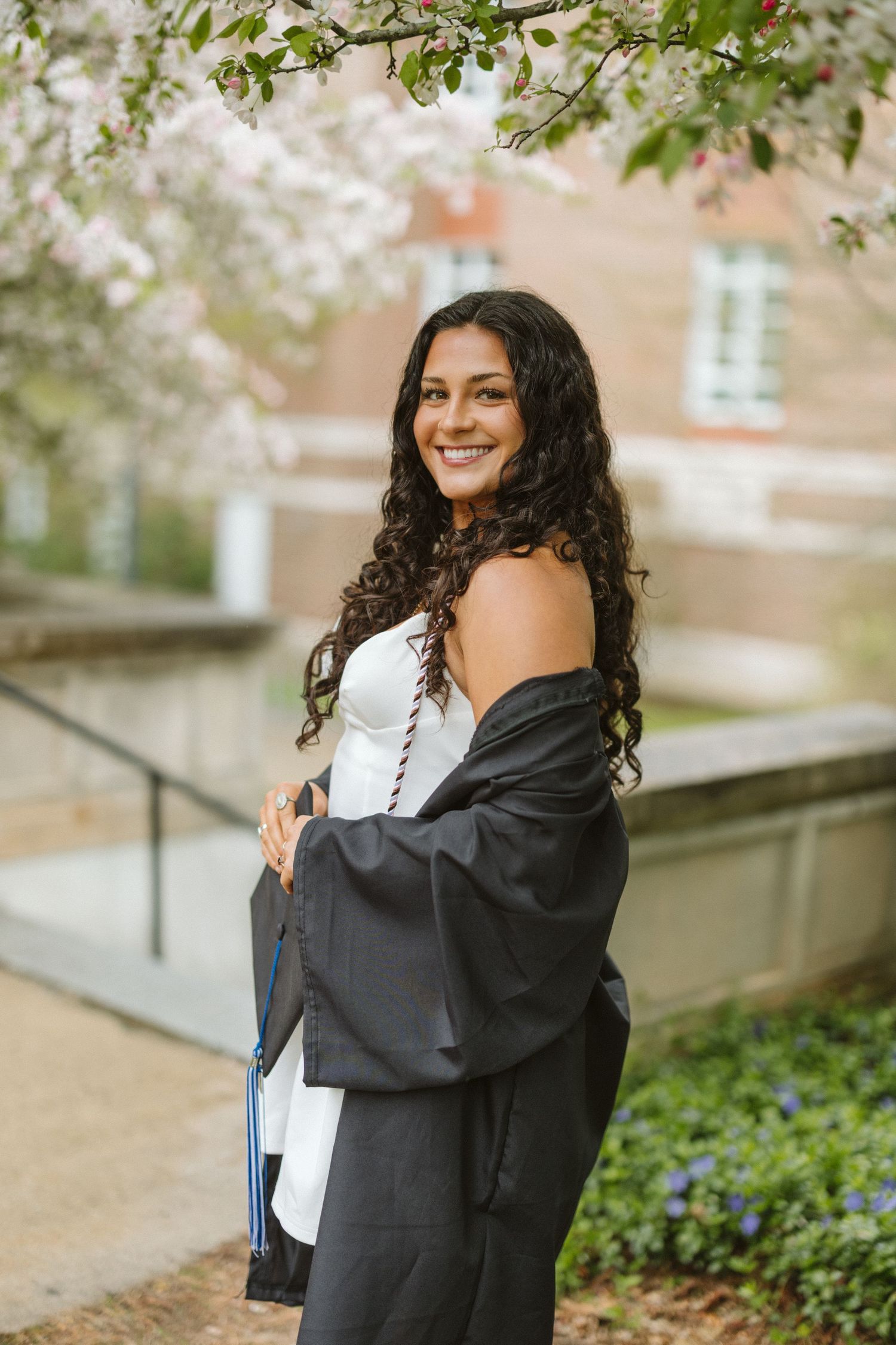 Smiling graduate in black gown holding cap under blooming cherry blossoms on a college campus in spring.
