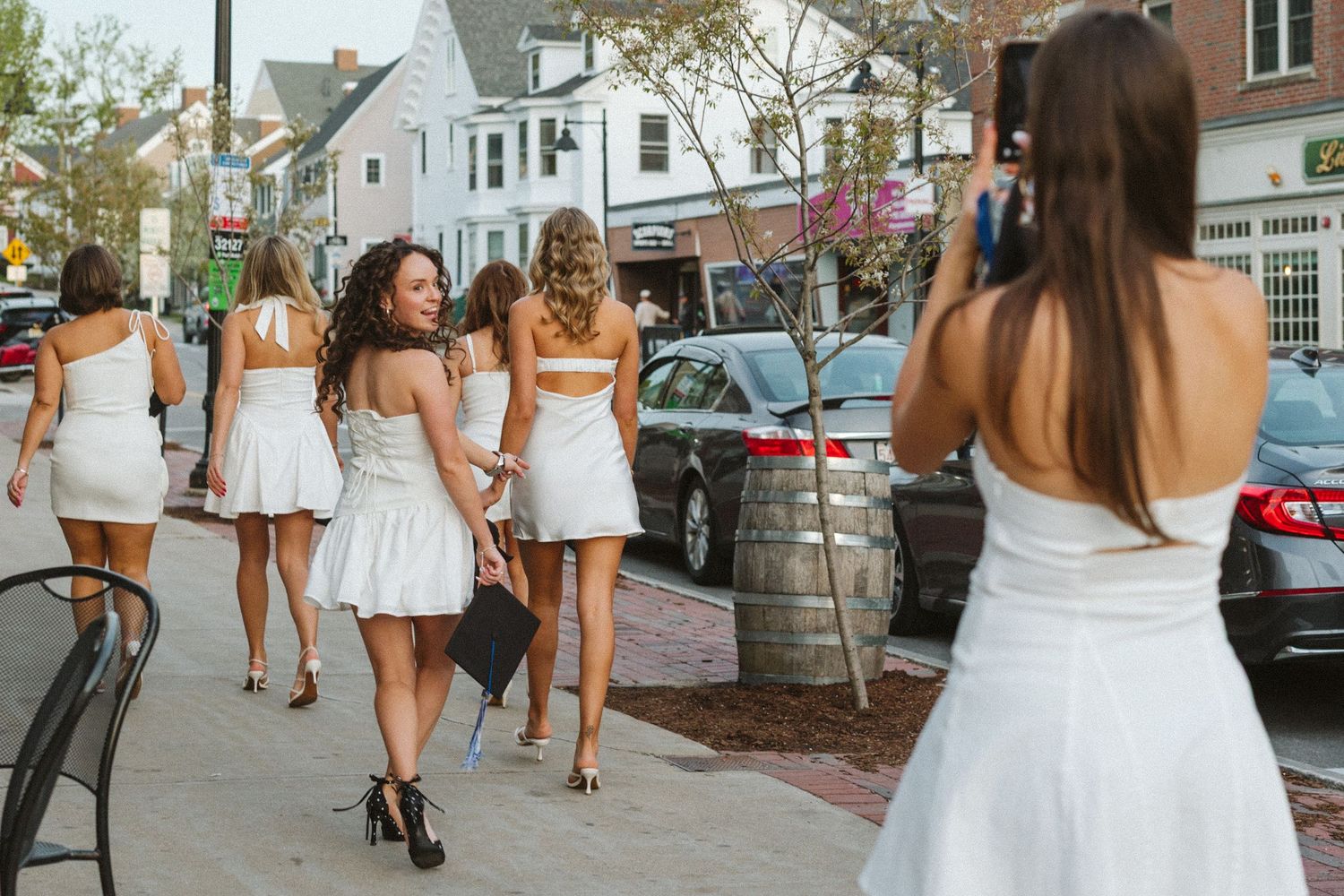 Group of women in white dresses walking down a sunny street while one photographs them from behind.