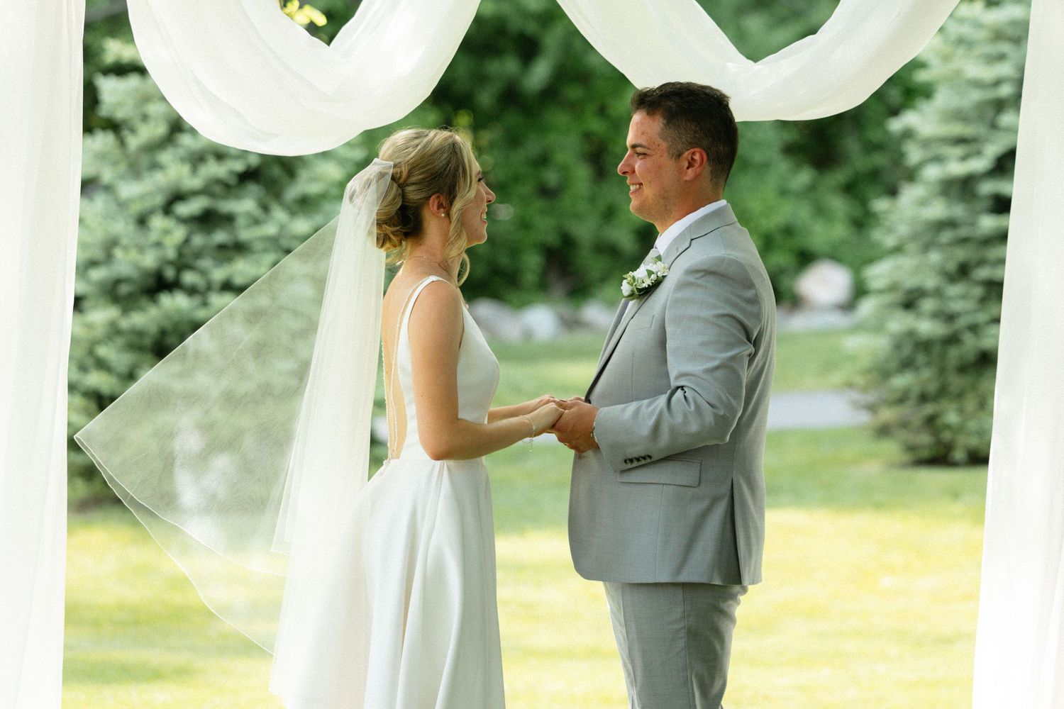 Bride and groom exchange vows outdoors under white draped arch surrounded by lush green trees at elegant garden wedding.